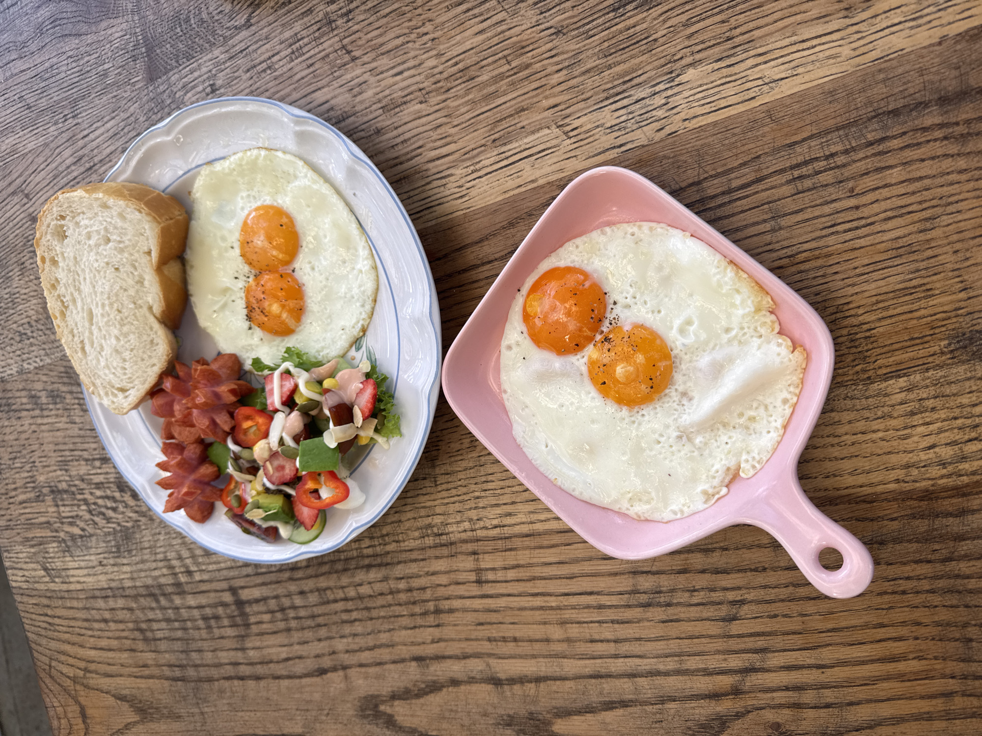 Two plates of food are pictured on a wooden table. The upper plate contains a slice of white bread, two fried eggs, sliced sausages, and a small salad with strawberries, cucumber, bell pepper, and greens. The lower plate, a pink dish with a handle, holds two fried eggs. Both plates are on a weathered wooden surface. The scene appears to be a breakfast setting, likely at a restaurant in Da Lat, Vietnam.