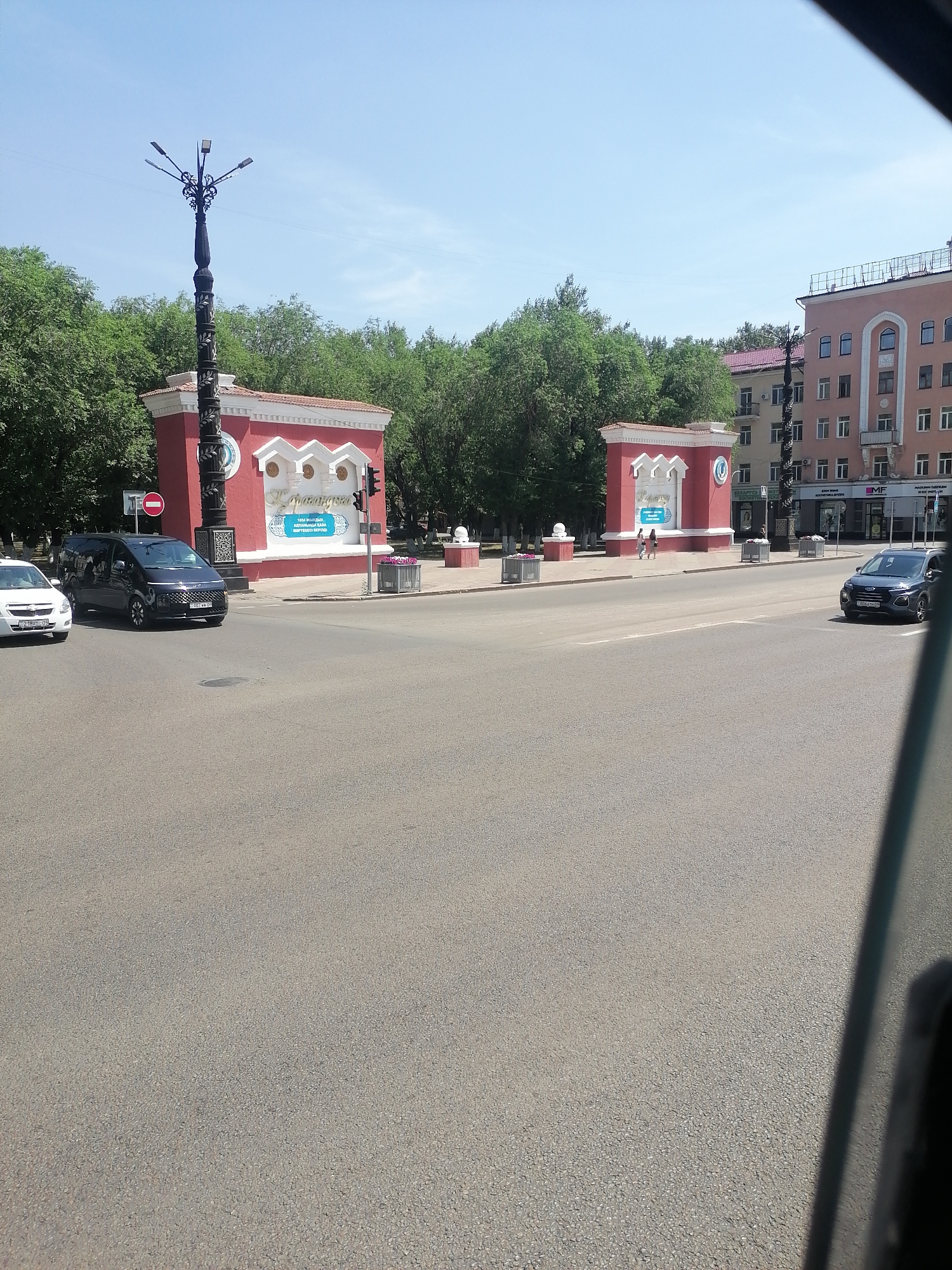 In a city scene, a street with asphalt pavement is visible. On the left side of the street, a white car is parked. Further down the street, a black minivan is driving, and to its right, a dark colored car is also in motion. In the background, there is a red structure with white detailing, possibly an entrance or monument, with ornamental lamps on top. Beyond this, trees line the area. To the right, a multi-story building with a light pink facade stands tall. The sky above is clear and blue.