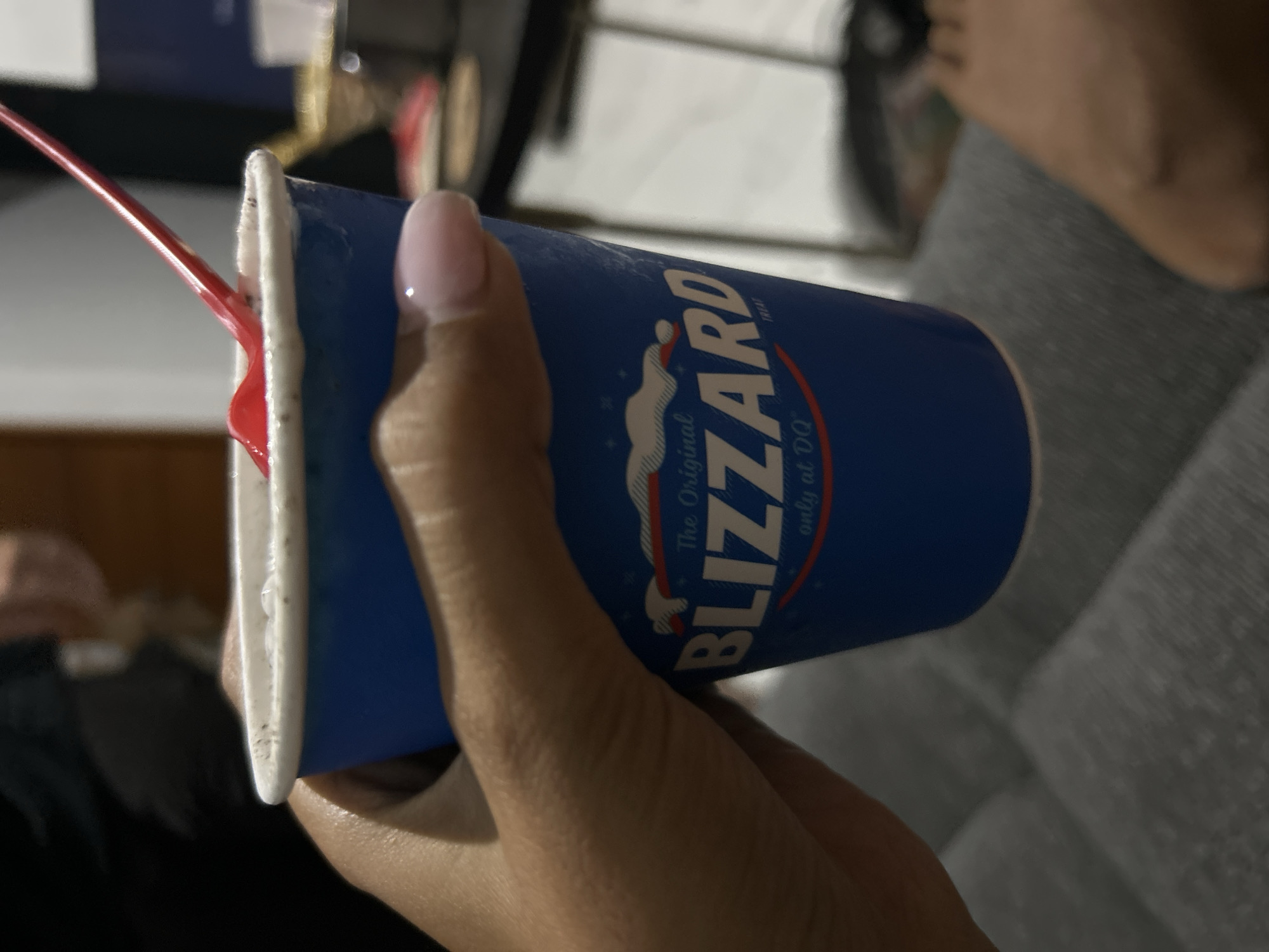 A close-up shot shows a person's hand holding a blue Dairy Queen Blizzard cup with a red spoon protruding from it. The cup has the Dairy Queen logo and "Blizzard" printed on it. The background is blurred, but appears to be an indoor setting with some furniture visible. The image is taken from a low angle, focusing on the cup and the hand. The person's fingernails are painted light pink. The environment suggests it is a casual setting, possibly a home or a restaurant. There is no information about the location in Sukawati, Indonesia.