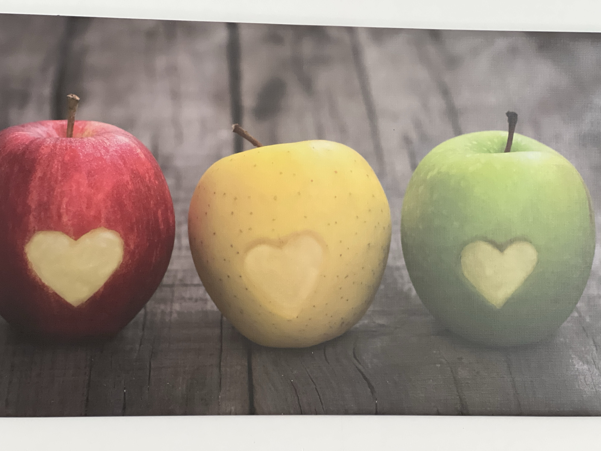 The image displays three apples: one red, one yellow, and one green, arranged horizontally on a wooden surface. Each apple has a heart shape carved into its surface. The background consists of a textured, greyish wooden surface. The image appears to be a photograph or a printed artwork, as indicated by the white border at the top and bottom. There is no identifiable information within the image that places it in Amberg, Germany.