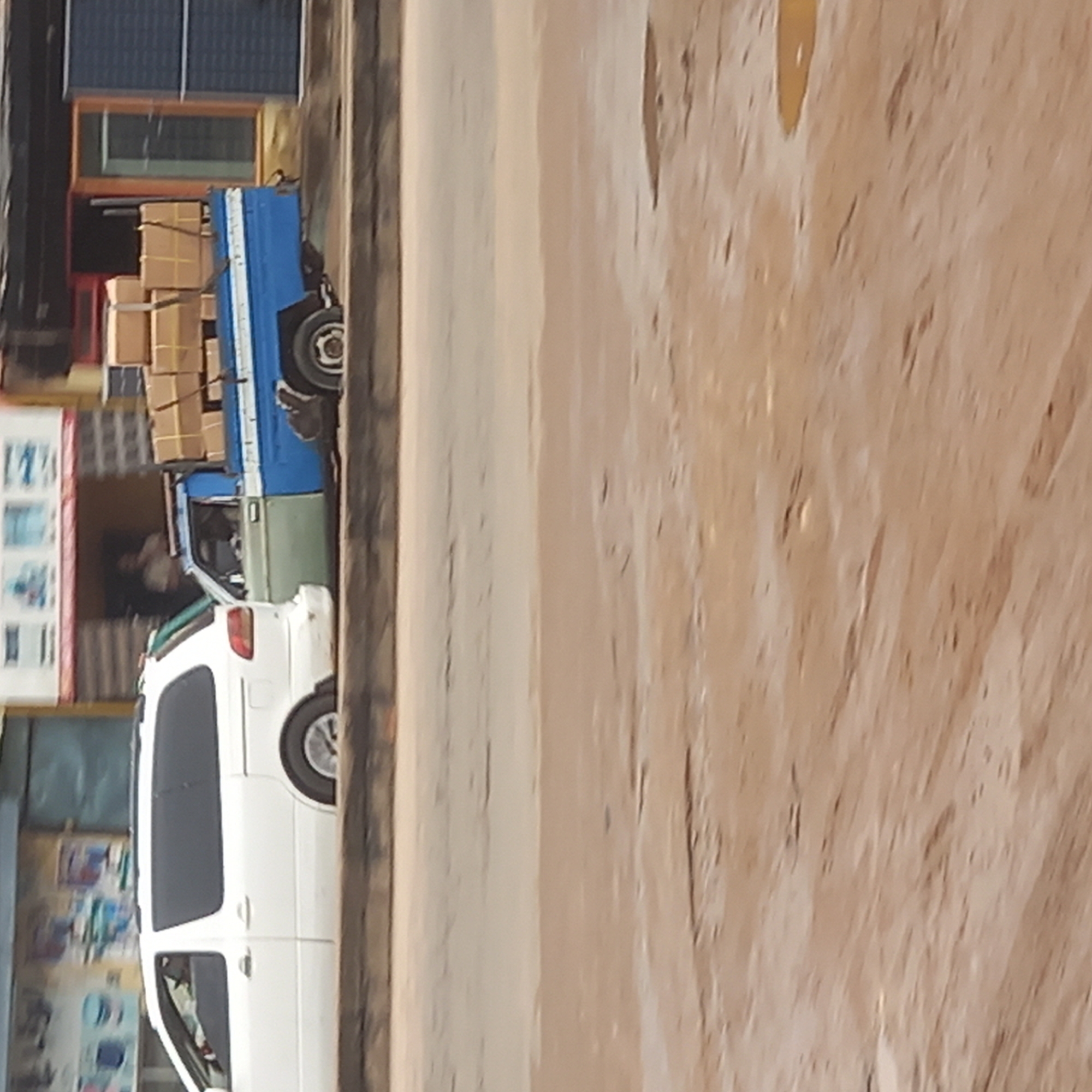 A white van and a blue truck loaded with cardboard boxes are parked on the side of a road in the city of Dadi Riba, Nigeria. A person is sitting inside the truck. A building with advertisements is beside the vehicles. The road is paved, and there is a dirt area beside it.