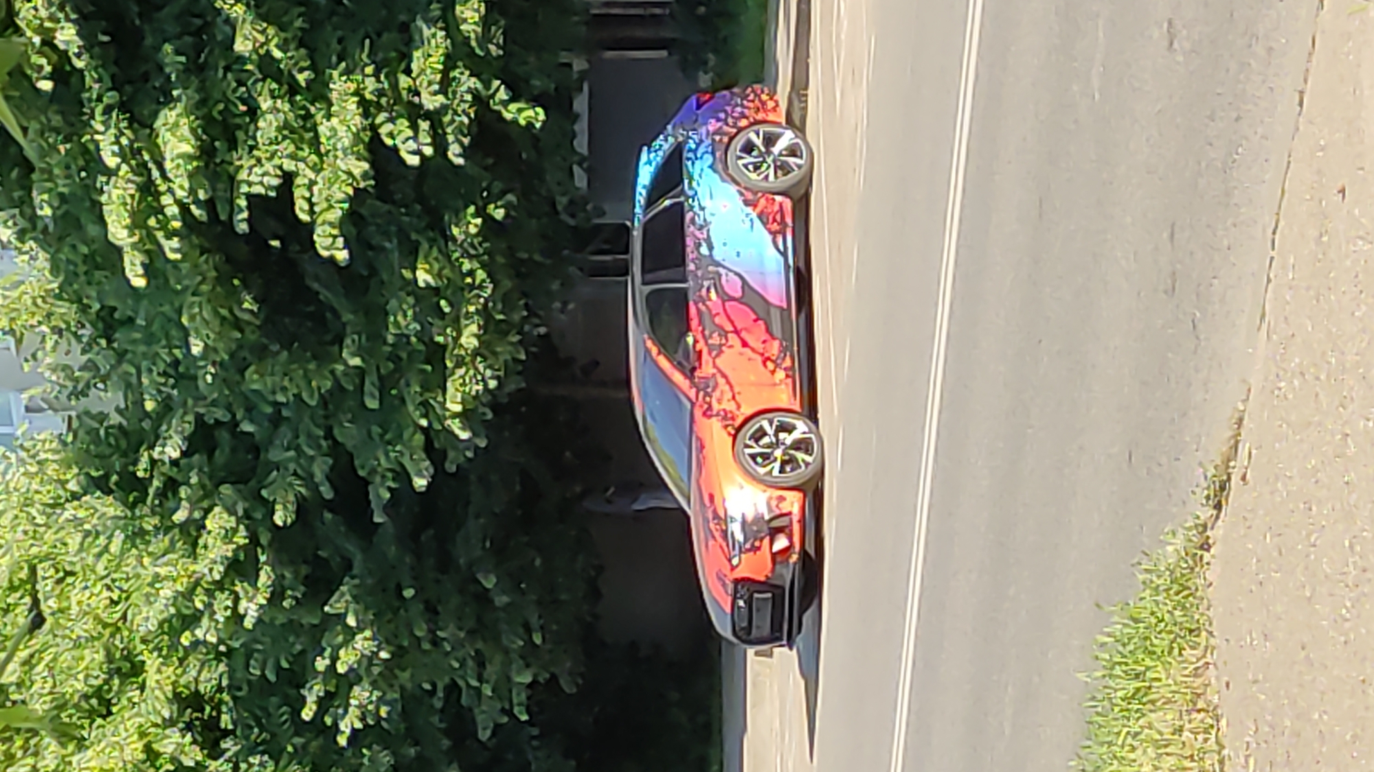 The image displays a custom-wrapped car parked alongside a road, bathed in bright daylight in Krasnodar, Russia. The main subject is a sedan featuring a vibrant, abstract design or wrap on its exterior, characterized by a dynamic blend of red, orange, blue, purple, and black patterns. The car is equipped with multi-spoke alloy wheels.
To the left of the car, a dense mass of lush, bright green foliage from trees or bushes fills a significant portion of the frame, indicating a natural or landscaped environment beside the road. The car itself is positioned on a paved surface, possibly a shoulder or parking area, which borders a road with clear double white lines visible. Further to the right, beyond the road markings, a strip of green grass grows alongside the paved surface.
The scene appears to be set on a clear, sunny day, with strong natural lighting casting subtle shadows. There are no people visible in the image, nor is there any discernible text on the car or in its surroundings. The car is stationary, with no specific activity taking place.