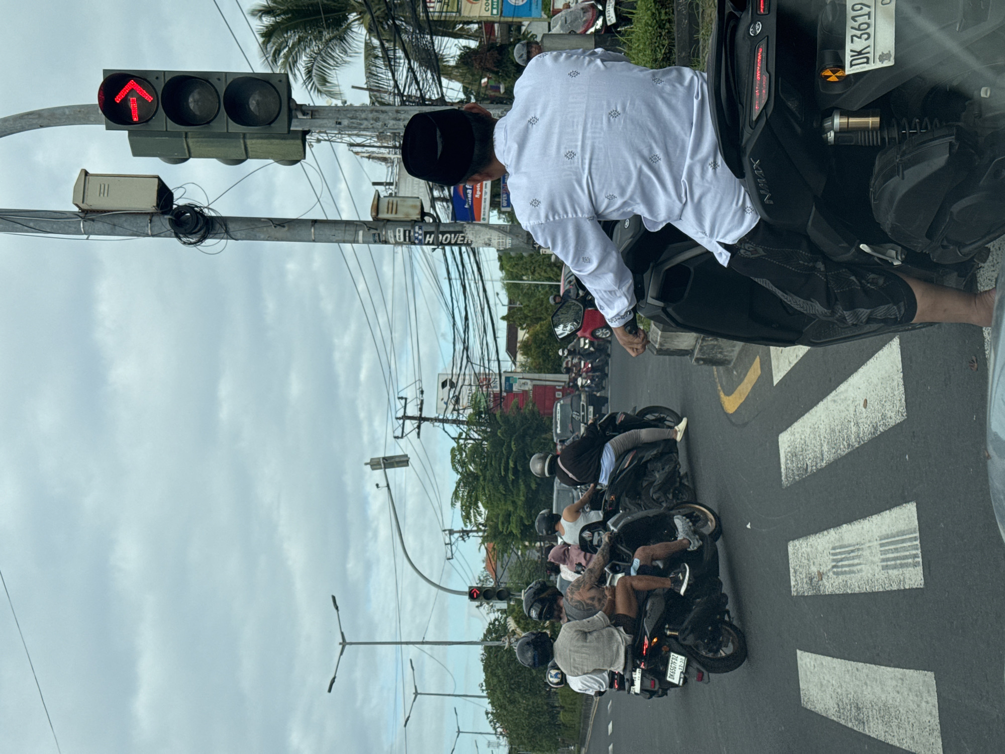 This media file captures a street scene in Kuta Selatan, Indonesia, likely during the day given the lighting. The sky is overcast with clouds. The primary subjects are a line of motorcyclists waiting at a traffic light. The traffic light is red, indicated by the illuminated red arrow pointing upwards.
In the foreground, a person wearing a white patterned shirt and a black peci (a cap commonly worn by Muslim men) is on a motorcycle. They are looking downwards, possibly at the traffic light or the road ahead. Behind them, several other motorcyclists are visible, most wearing helmets. The motorcycles are lined up on a paved road with white pedestrian crossing markings.
Other details include numerous electrical wires and poles, typical of urban Southeast Asian environments, and lush green trees in the background. Some signage is visible on poles, though it is mostly illegible. The overall impression is of a busy intersection with traffic waiting for the signal to change. The mood seems neutral, as is typical for a traffic stop. Visible text includes the license plate "DK 3619 ON" on one of the motorcycles.
