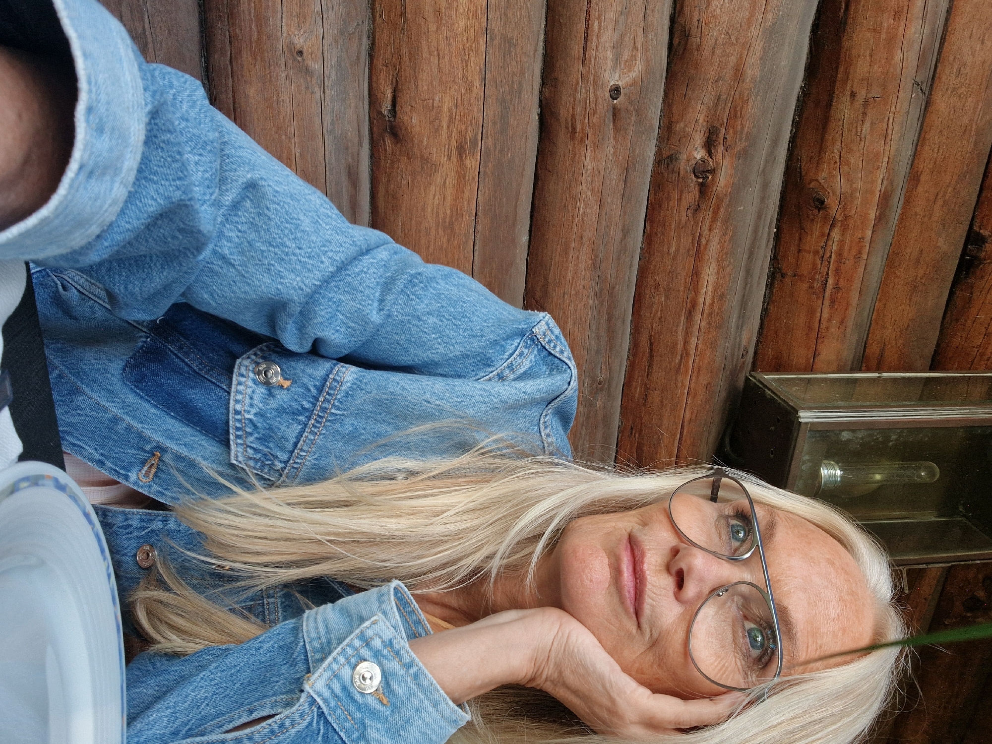 A medium shot shows a woman with long, blonde hair wearing glasses and a denim jacket. She is looking upwards and to the right, with her right hand resting on her chin. She is positioned in front of a wooden wall, and to her left, a lantern is visible hanging on the wall. A white plate with blue and white patterned trim is in the foreground. The image appears to be taken outdoors, possibly at a cafe or restaurant with a rustic aesthetic. The location is Nynäshamn, Sweden.