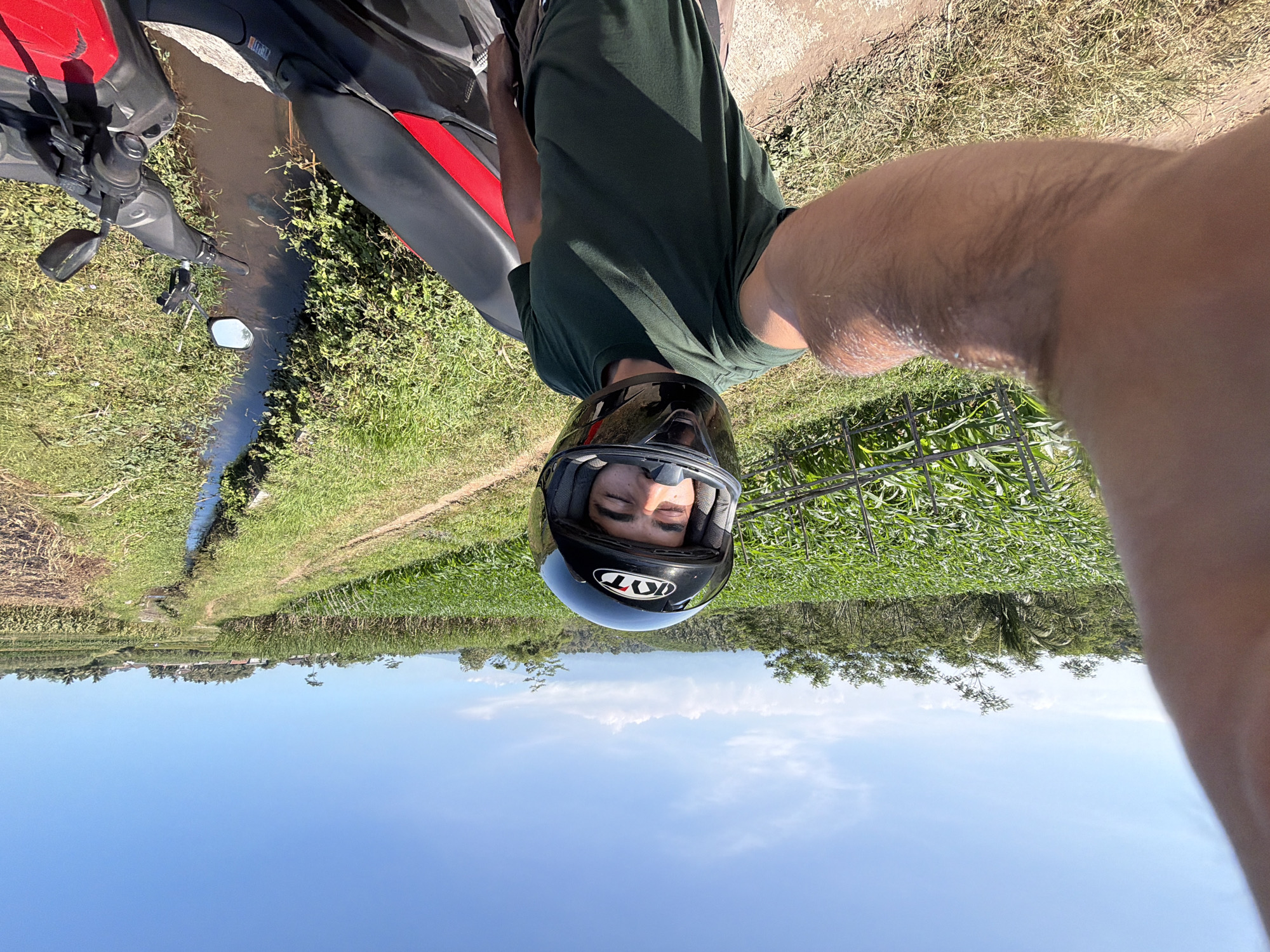 A young man wearing a green t-shirt and a black motorcycle helmet with "KYT" visible on it, is taking a selfie. He is sitting on a red and black motorcycle next to a small stream with green grass and some cultivated fields. The sky is bright blue with a few white clouds, suggesting it is daytime and the weather is clear. The scene appears to be rural, likely in Indonesia given the vegetation and the presence of a motorbike. The man's eyes are closed, and he has a relaxed expression, suggesting he is enjoying the moment or perhaps resting. In the background, there are trees and a distant landscape that suggests rolling hills. The overall mood is peaceful and serene, capturing a moment of leisure in a natural setting.