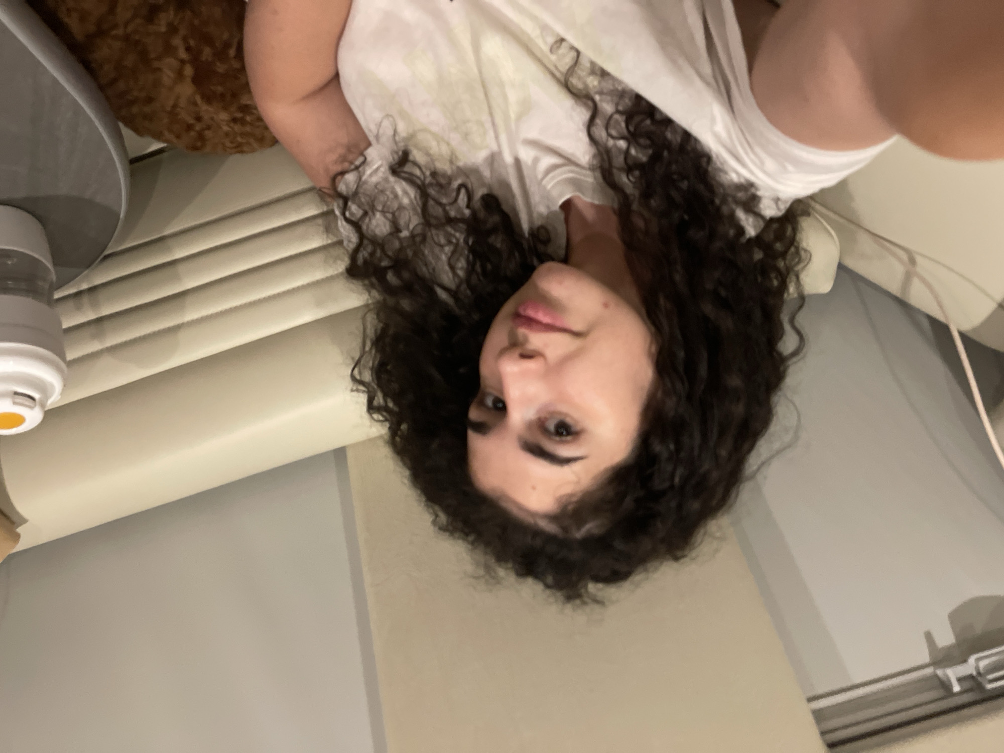 The scene depicts a young woman in an indoor setting, located in Koprinka, Bulgaria. She has dark, curly hair and fair skin, with dark eyes and defined eyebrows. She is wearing a light-colored t-shirt, possibly with a subtle patterned design, and is looking directly at the camera with a slight, closed-lip smile. A small blemish is visible below her lower lip on the right side of her face.
She is seated on a light-colored, possibly cream or beige, upholstered sofa or bench featuring horizontal ribbed stitching on its backrest. To her lower right, a portion of a reddish-brown, curly-haired dog is visible, resting on the seating. To her immediate right, there is a small, round, gray table on which a white and clear portable beverage container with a yellow cap is placed. The background consists of light-colored walls or paneling. On the far left, a metallic handle or latch mechanism is partially visible on what appears to be a doorframe or an internal partition, suggesting a structured interior such as a private jet, RV, or similar specialized compartment. The lighting is soft and ambient.