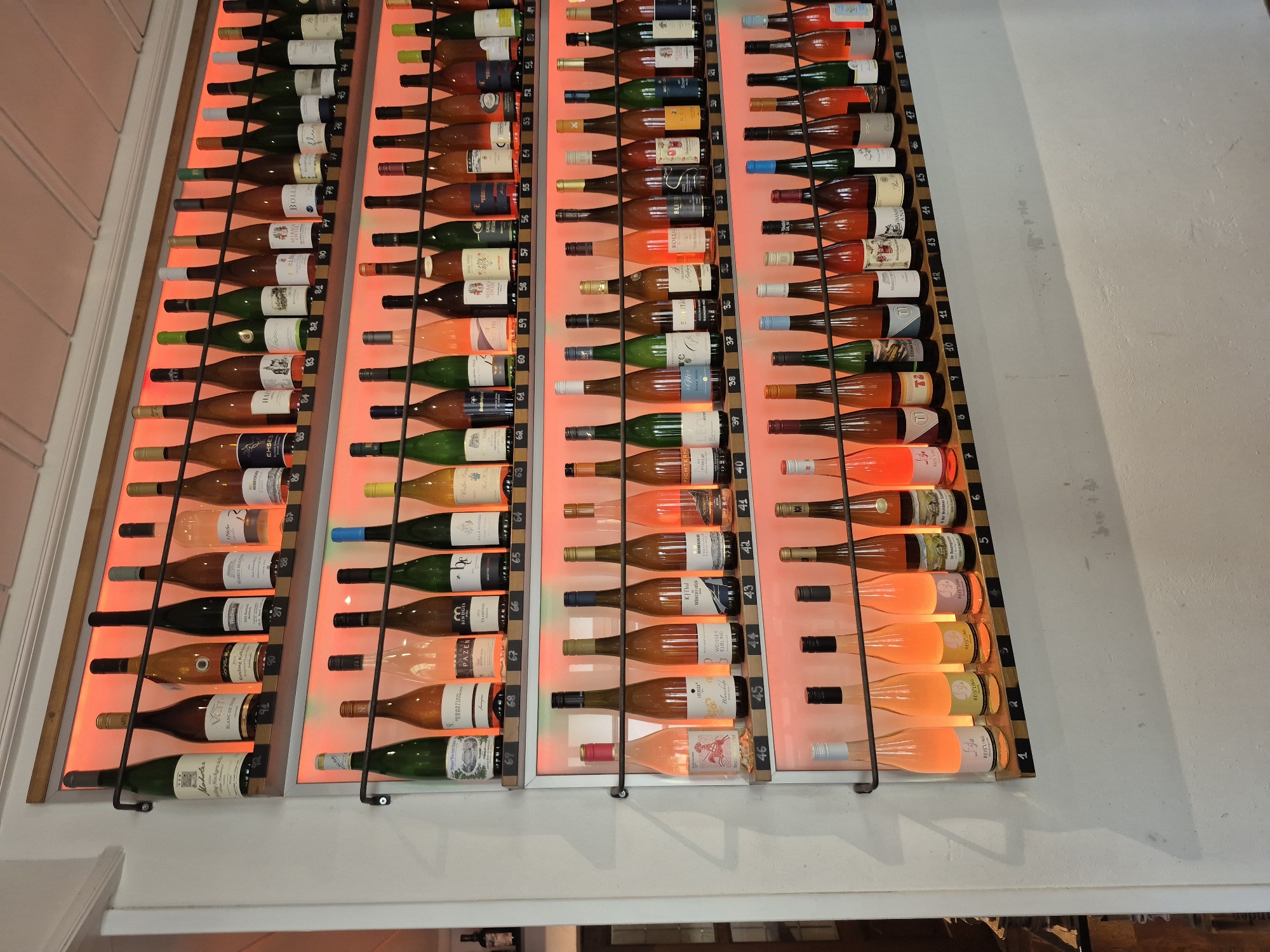 This image captures a wall-mounted wine display within a commercial establishment in Bernkastel-Kues, Germany. The display consists of six horizontal shelves, each supported by two parallel metal rods, against a plain white wall. Each shelf is backlit with a warm, reddish-orange light that illuminates the wine bottles and enhances the visibility of their contents.
A total of 92 wine bottles are displayed, sequentially numbered on small, light-colored wooden blocks positioned directly beneath each bottle. The numbering begins with '1' on the bottom-right of the lowest shelf and ascends to '92' on the top-left of the highest shelf.
The bottle count per shelf, from top to bottom, is as follows:
* Top shelf: 9 bottles (numbered 92 to 84)
* Second shelf: 18 bottles (numbered 83 to 66)
* Third shelf: 18 bottles (numbered 65 to 48)
* Fourth shelf: 18 bottles (numbered 47 to 30)
* Fifth shelf: 18 bottles (numbered 29 to 12)
* Bottom shelf: 11 bottles (numbered 11 to 1)
The bottles vary in glass color, including dark green, dark brown, and clear, and contain wines exhibiting a spectrum of colors from deep golden amber to various shades of pink (rosé) and light yellow. Many labels are visible, displaying German text and identifiable vintner names such as 'Müller Thurgau,' 'Weber,' 'Markus Molitor,' 'PaZen,' 'Sebastian Erbeldinger,' 'Leitsch,' 'Bollig,' 'Kitzmuhle,' and 'Pichter.' These labels indicate the German origin of the wines, typical of the Bernkastel-Kues region.
The surrounding environment includes a clean, white wall surface. Above the display, a white ceiling with visible paneling is present. To the far left, a vertical white architectural element, likely a door frame or wall corner, is visible. A dark, out-of-focus wooden object, possibly a shelf or cabinet, is partially visible in the lower left background. No individuals are present in the image.