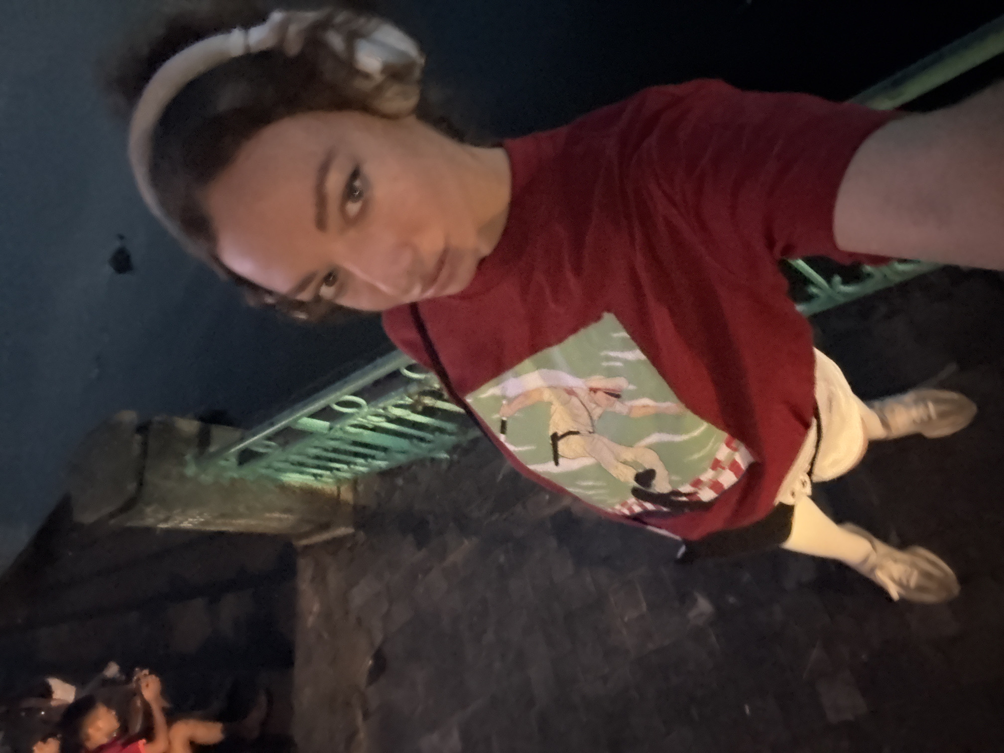 A person wearing headphones and a red t-shirt with a graphic is standing on a stone walkway next to a railing. The person is looking at the camera and is in the foreground. In the background, other people are sitting and looking at phones. The scene appears to be outdoors at night. The location is not Hà Nội, Vietnam.