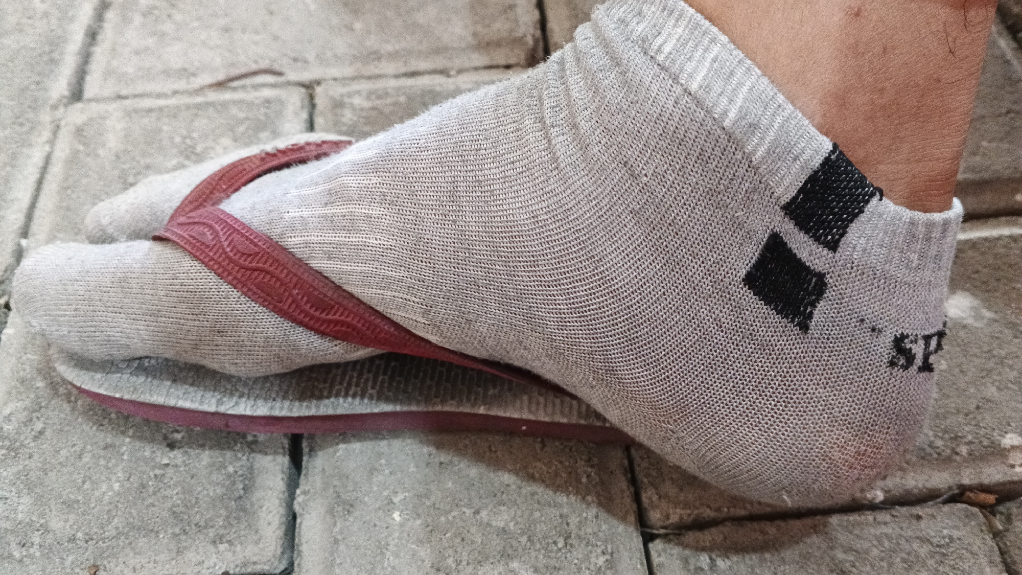 A close-up, side-view shot focuses on a person's left foot, clad in a light gray ankle sock and a red flip-flop. The sock has a distinct weave pattern and features two small black diamond shapes near the ankle, along with the letters "SP" printed on the heel. The flip-flop's red straps pass between the sock-clad big toe and the second toe, and its sole rests on a textured, gray, paver-stone surface, common in outdoor pedestrian areas of Yogyakarta, Indonesia. The lighting suggests an outdoor environment, possibly during daytime, with shadows hinting at the time of day.