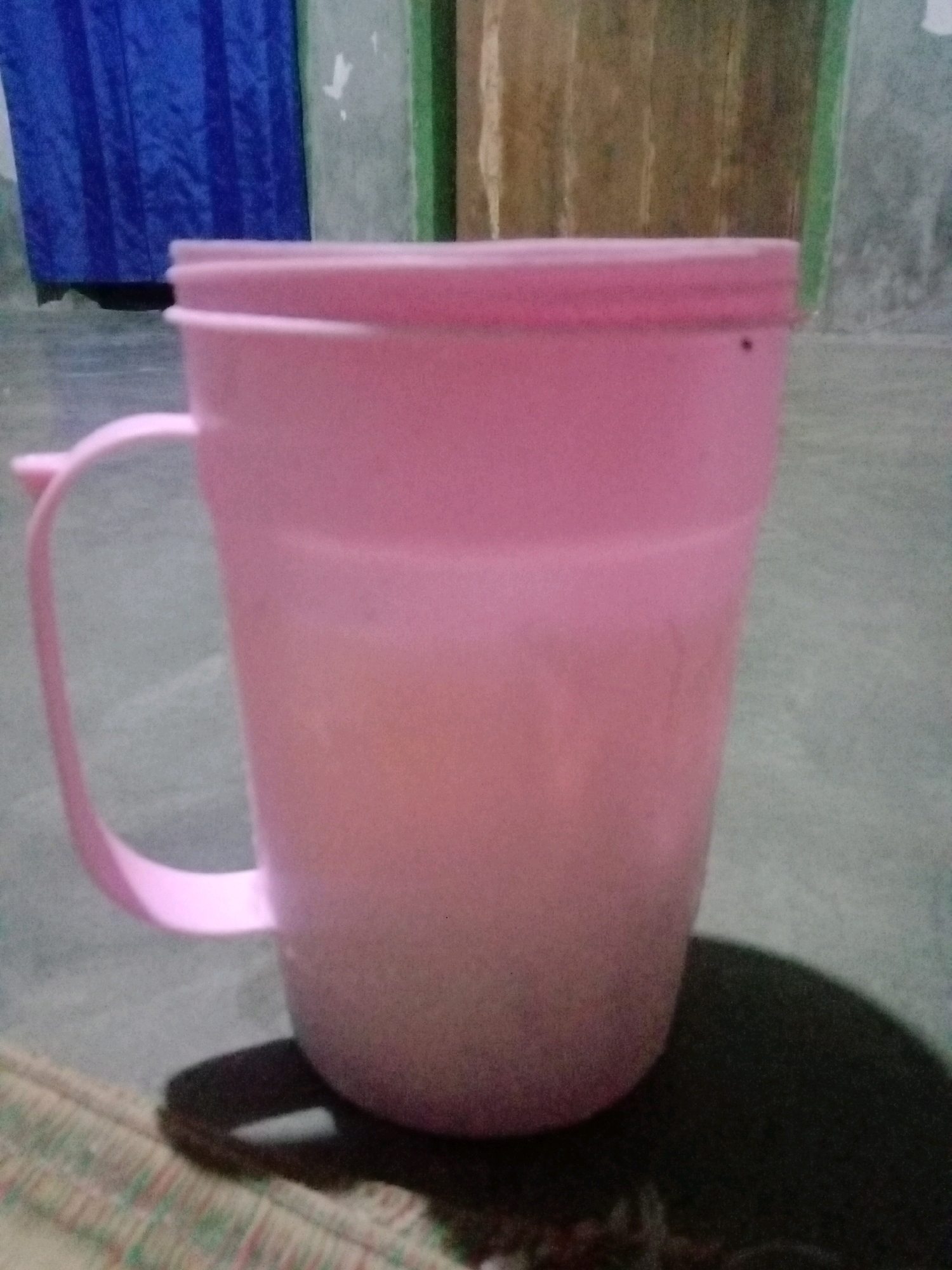 A pink plastic mug with a handle is on a woven mat. The mug is filled with a light-colored liquid. In the background, there is a blue curtain and a beige wall with a green vertical stripe. The mug is casting a shadow on the mat. The scene is indoors in Utama Jaya, Mataram, Indonesia.