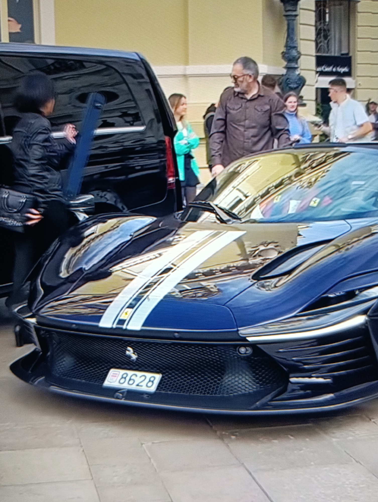 The image shows the front of a blue Ferrari Daytona SP3 with white racing stripes on the hood. The car has a Monégasque license plate with the number "8628." To the left of the car is a black SUV, and a woman in a black leather jacket appears to be interacting with someone inside the SUV. Behind the car and SUV, a group of people are standing on a paved area. Among them is a man wearing glasses and a brown shirt. In the background, there is a building with a black sign that reads "Un Ciel d'Anges." The location appears to be Monaco, indicated by the car's license plate.