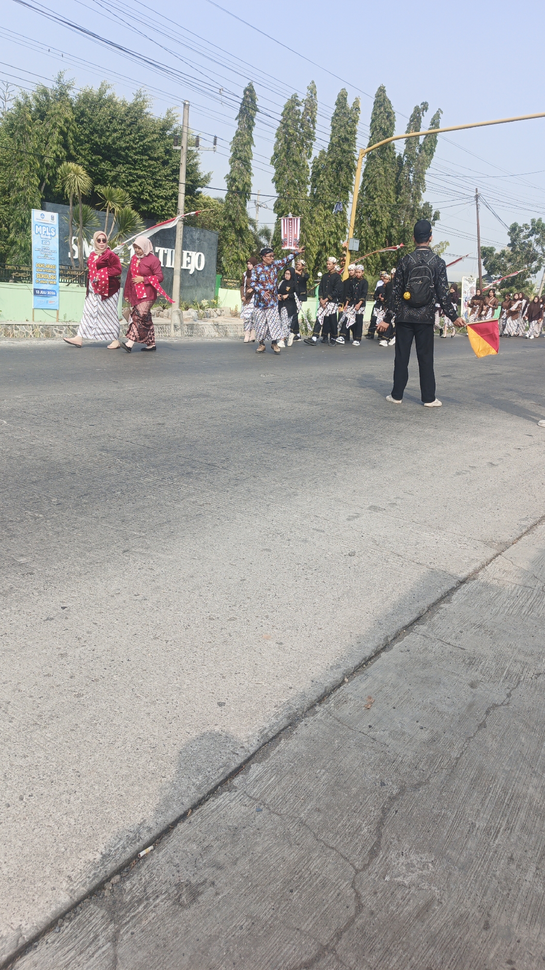 In the city of Sumengko, Indonesia, a procession of individuals is captured on an asphalt road under a clear sky. The scene depicts a group of students, some in traditional Indonesian attire including batik shirts, sarongs, and headwear, walking in formation. A few individuals are seen holding banners or flags, one of which is a red and yellow triangular flag being held by a person wearing a black backpack. Behind the students, a building with the inscription "SMK TI LEJO" is visible, suggesting the location might be a school or associated with a school. The environment includes trees lining the background and a sidewalk with a light pole. The road exhibits some wear and tear, with visible cracks and a discarded cigarette butt.