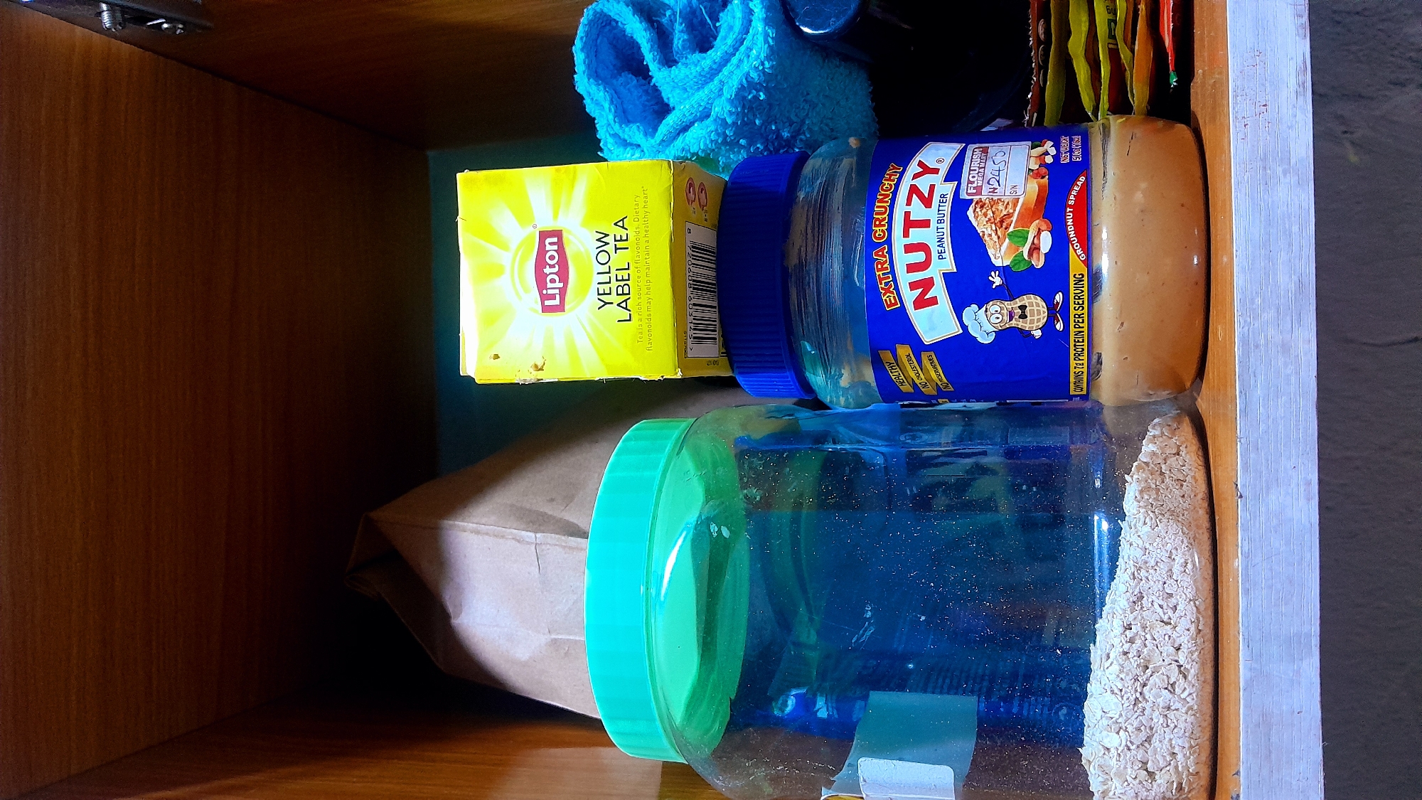 This media file captures a close-up, rotated view inside a wooden cupboard or shelf, likely in a kitchen or pantry in Umuigbekwo, Nigeria. The image is oriented sideways, with objects appearing to lie on their sides.
The main subjects are various food items and a cleaning cloth. Prominently featured is a bright yellow box of Lipton Yellow Label Tea, which shows some wear. Text on the box includes "Lipton", "YELLOW LABEL TEA", and a health claim: "Tea is a rich source of flavonoids. Dietary flavonoids may help maintain a healthy heart."
Next to the tea box is a blue-lidded jar of "NUTZY EXTRA CRUNCHY PEANUT BUTTER." The label specifies "PEANUT BUTTER," "GROUNDNUT SPREAD," and "CONTAINS 7g PROTEIN PER SERVING." Other notable text on the label includes "FLOURISH MEGA MAN," "N2450," "S/N," "HEALTHY," "NO CHOLESTEROL," and "NO PRESERVATIVES." The "N2450" likely indicates a price in Nigerian Naira, reinforcing the specified location context.
Below the peanut butter jar is a large clear plastic jar with a bright green lid, filled with what appears to be oatmeal or rolled oats. Behind the tea and peanut butter, a crumpled blue microfiber towel or cloth is visible. In the lower left, part of a brown paper bag is tucked away, and behind the items on the right, several small, colorful packets or sachets can be seen.
The setting is dimly lit, suggesting an interior space away from direct light, with the wooden surfaces of the cupboard creating a warm, enclosed environment. No specific activity or event is taking place; the image is a still life of stored provisions.