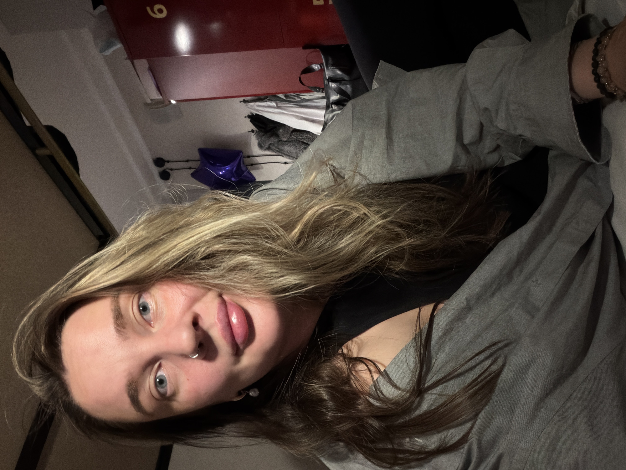 The image shows a young woman in an indoor setting, likely in Moscow, Russia. She has long, wavy hair, a mix of blonde highlights and darker brown tones, and striking blue eyes. Her face is angled towards the camera, with a direct and slightly playful expression, her lips parted to reveal her tongue. A silver septum piercing adorns her nose. She is wearing a dark top beneath a loose-fitting, light gray shirt or jacket. On her visible right wrist, she wears several beaded bracelets, one dark and one lighter in color.
The background is dimly lit, suggesting evening or night. To her left, a red locker or cabinet is partially visible, clearly marked with the yellow-orange number "6." Below it, a bright purple foil balloon rests on a surface. Other objects, including what appears to be a black bag or folded fabric, are indistinctly visible in the cluttered background, indicative of a casual, personal space. The scene conveys an informal and intimate atmosphere.
