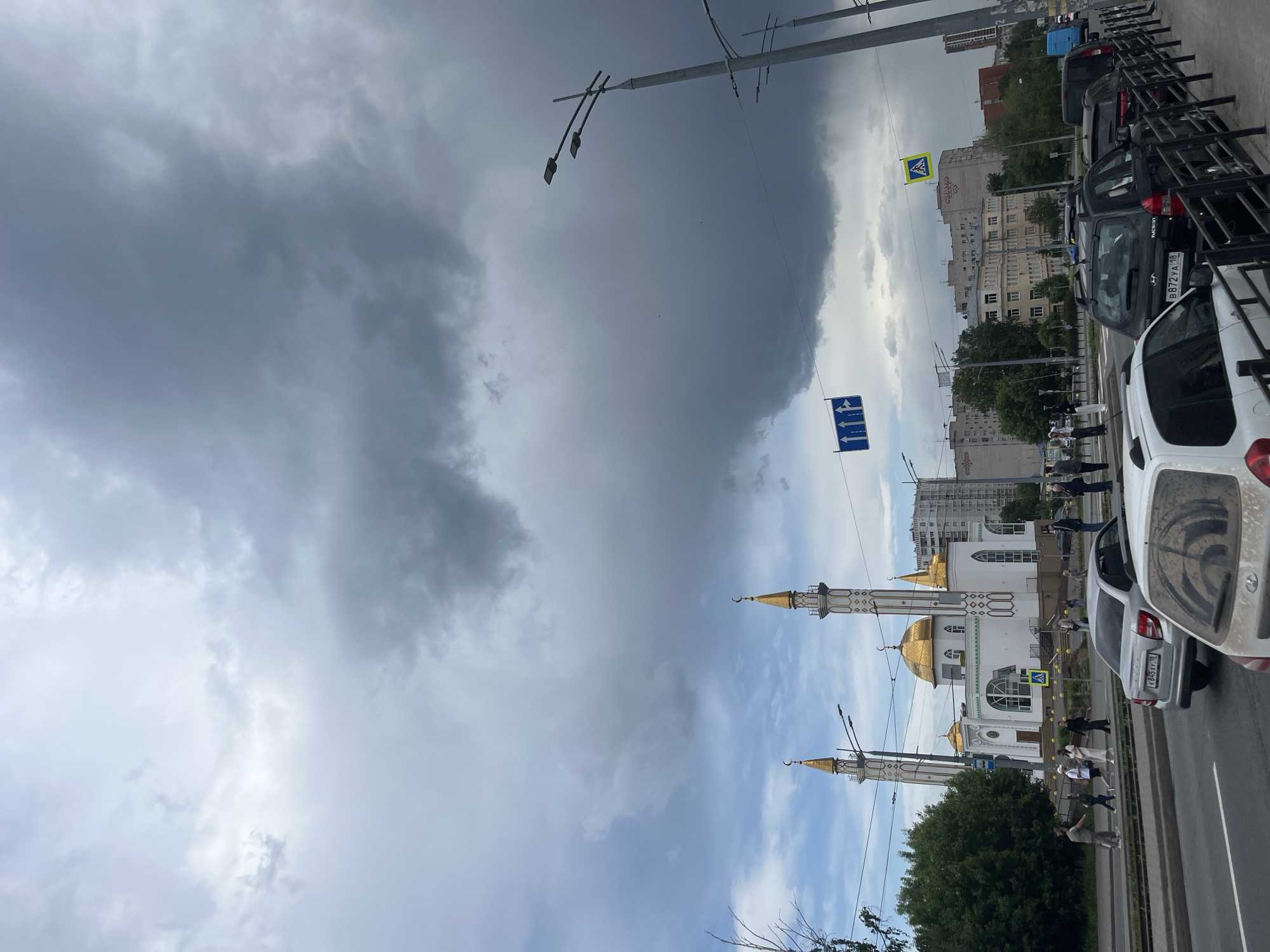 A street view in Izhevsk, Russia shows the sky filled with dark, overcast storm clouds above the white-walled mosque with golden domes and minarets. Cars are parked alongside the road with several pedestrians visible near the mosque. Buildings line the opposite side of the street beyond the mosque. Streetlights and traffic signs are present.