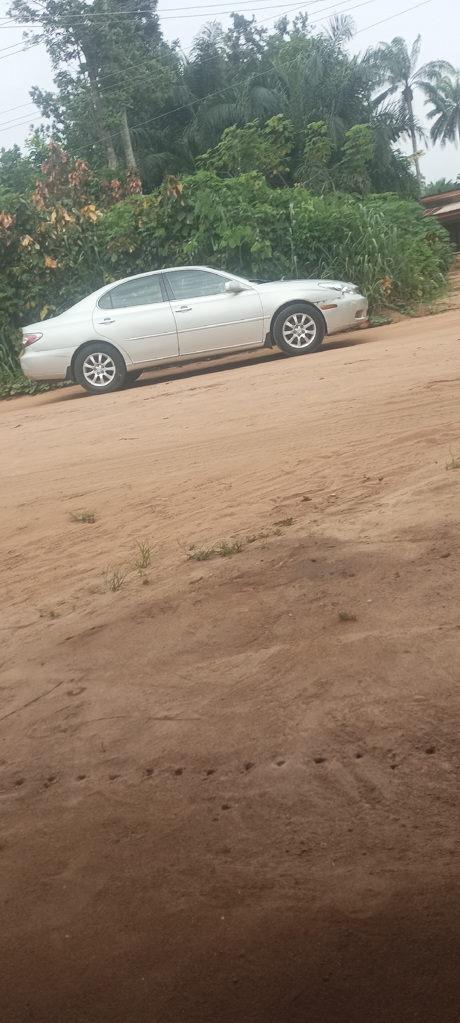 A silver sedan is parked on an unpaved, sandy road in what appears to be a rural or semi-rural location, possibly Amauzu, Nigeria. Lush, dense green foliage and trees form a backdrop. The road surface is uneven and shows signs of recent activity, with footprints or tracks visible. There are no visible people in the scene.