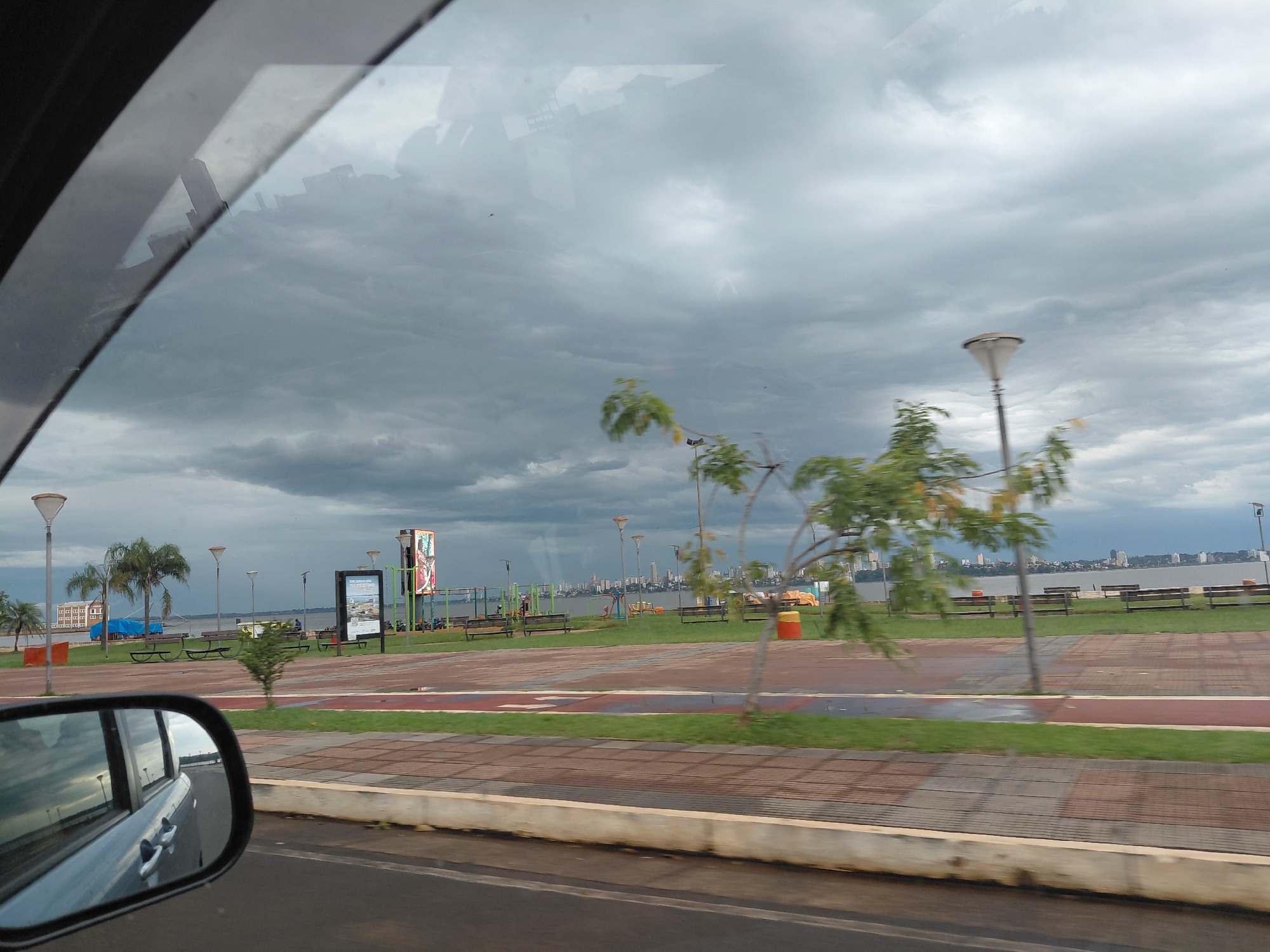 The image depicts a view of the Costanera de Encarnación in Paraguay from inside a moving vehicle. The sky is heavily overcast with dark clouds. In the foreground, the side mirror of the vehicle reflects the passing scenery, including parts of the vehicle's interior and the road. Beyond the road, a paved promenade with benches, grass, and trees stretches towards a waterfront. Several lampposts are visible along the promenade. In the distance, across a body of water, a city skyline with numerous buildings is visible. The overall scene suggests a cloudy, possibly humid day in Encarnación, with the Costanera serving as a recreational area.