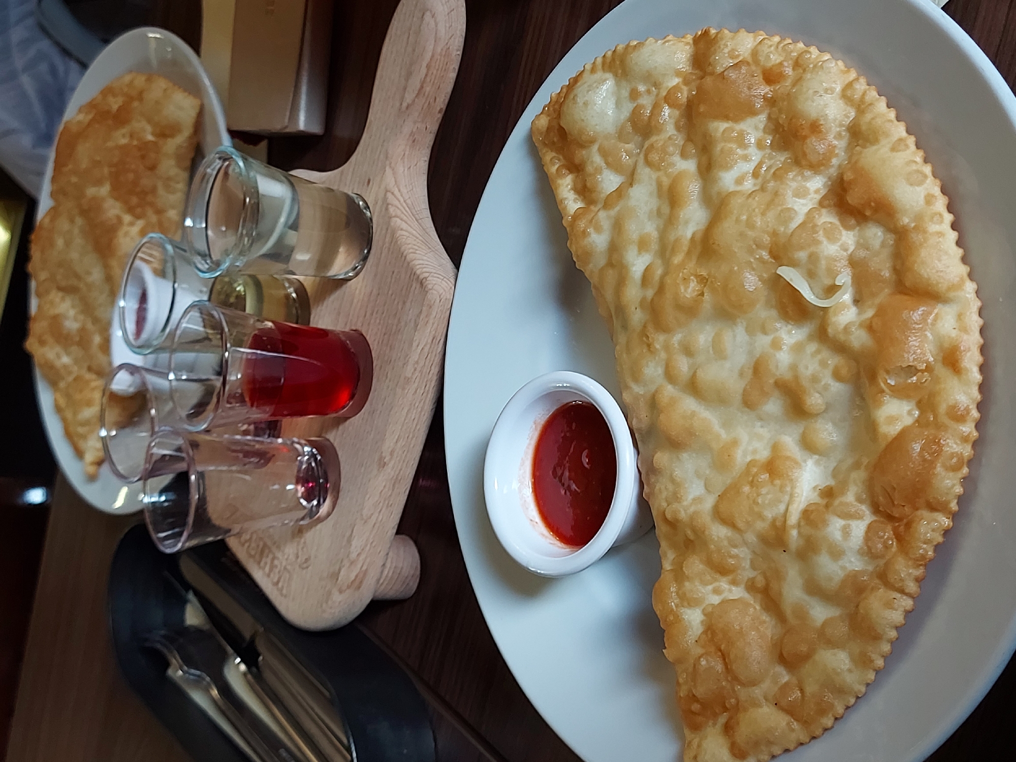 The image depicts a meal presentation, potentially in a restaurant or food establishment. A large, crescent-shaped fried pastry lies on a white plate accompanied by a small, white ramekin of red sauce. To the left, a wooden serving board supports several small shot glasses containing different liquids. A plate with additional fried pastries is positioned in the background. Cutlery is also visible at the bottom. The location is Saint Petersburg, Russia.