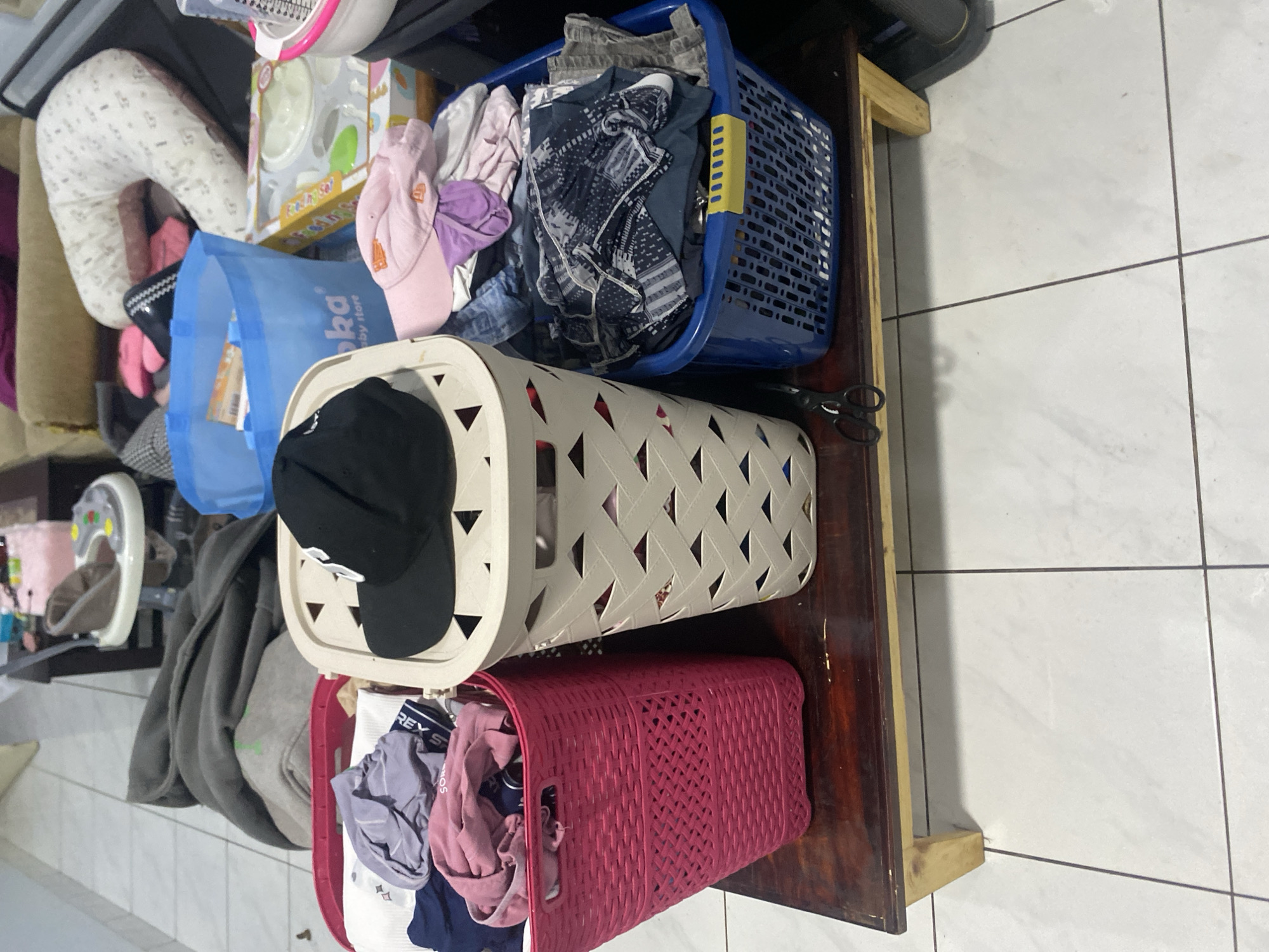 A close-up, elevated shot captures three laundry baskets overflowing with clothes, situated on a dark wooden shelf. The basket on the left is a vibrant pink, the middle one is cream-colored, and the one on the right is a deep blue. The cream basket prominently displays a black baseball cap with a white logo. The backdrop reveals a glimpse of a residential setting, possibly a living room, with a light-colored tiled floor and scattered household items. The overall impression is one of domestic clutter, likely in a home in Dukuh, Indonesia.