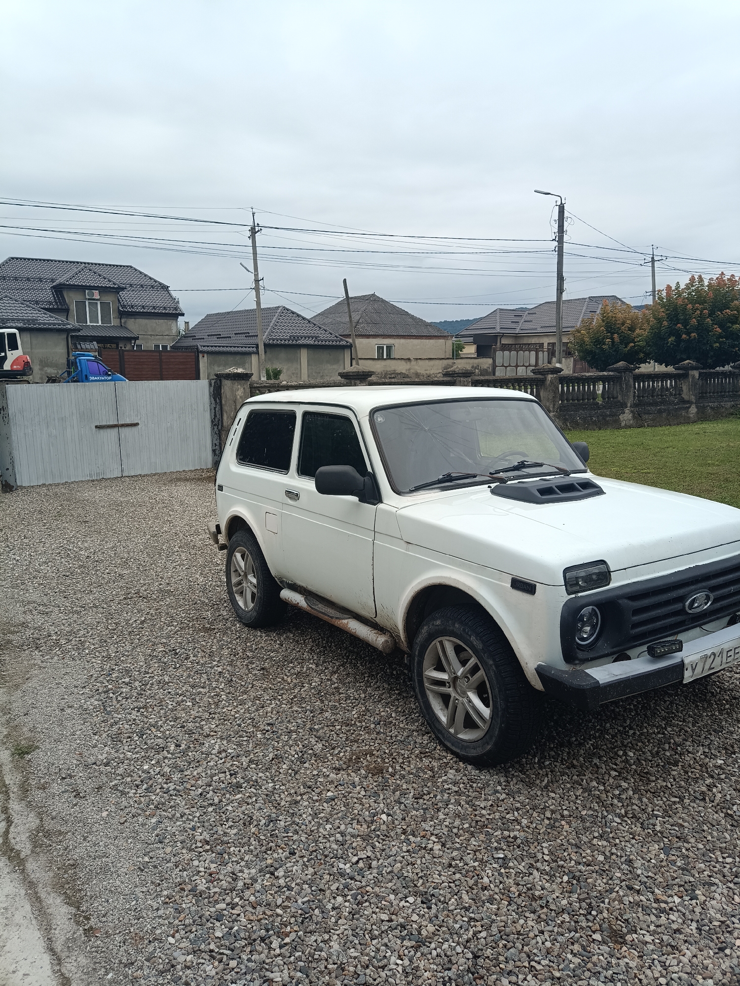 The image shows a white Lada Niva urban SUV parked on a gravel driveway in front of a residential property. The car is positioned at an angle, with its front-right side facing the viewer. The vehicle appears to be slightly dirty, with visible mud splatters on the lower body panels. The license plate is partially visible and reads "Y 721 EE".

Behind the car, a large, light-colored metal gate is closed. To the left of the gate, a portion of a white building or structure is visible, possibly a garage or storage area, with a red and white object protruding from its side. Further in the background, several residential houses with dark, sloped roofs are visible. Some of these houses have multiple windows and one has an air conditioning unit installed near the roofline.

The environment is overcast, with a cloudy sky. Power lines and utility poles are present in the background, crossing above the houses. A fence with decorative pillars runs along the edge of a grassy area to the right of the car. The ground is covered in a thick layer of gravel, with a concrete edge visible on the far left of the image. The overall impression is a rural or semi-urban residential setting. The specific location is identified as Kenzhe, Russia. No people are visible in the image.