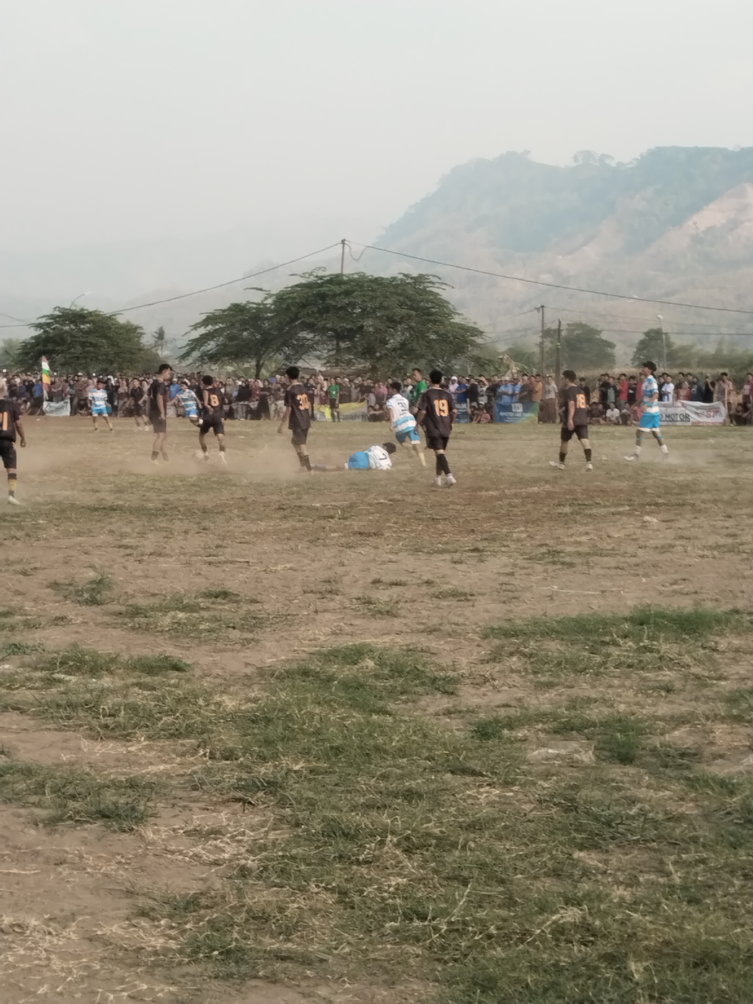 The image depicts a football match taking place on a dirt field with sparse grass, likely in Margasari, Indonesia, given the context. A crowd of spectators watches from the sidelines, some standing behind a banner. Several players in distinct team jerseys are actively engaged in the game. One player in a blue and white striped jersey is attempting to control the ball near the center of the field, kicking up dust. Other players, some in dark jerseys with numbers like 18, 19, and 30, are visible in various positions on the field. In the background, trees and hazy mountains form the landscape. The sky is overcast, contributing to the hazy atmosphere.
