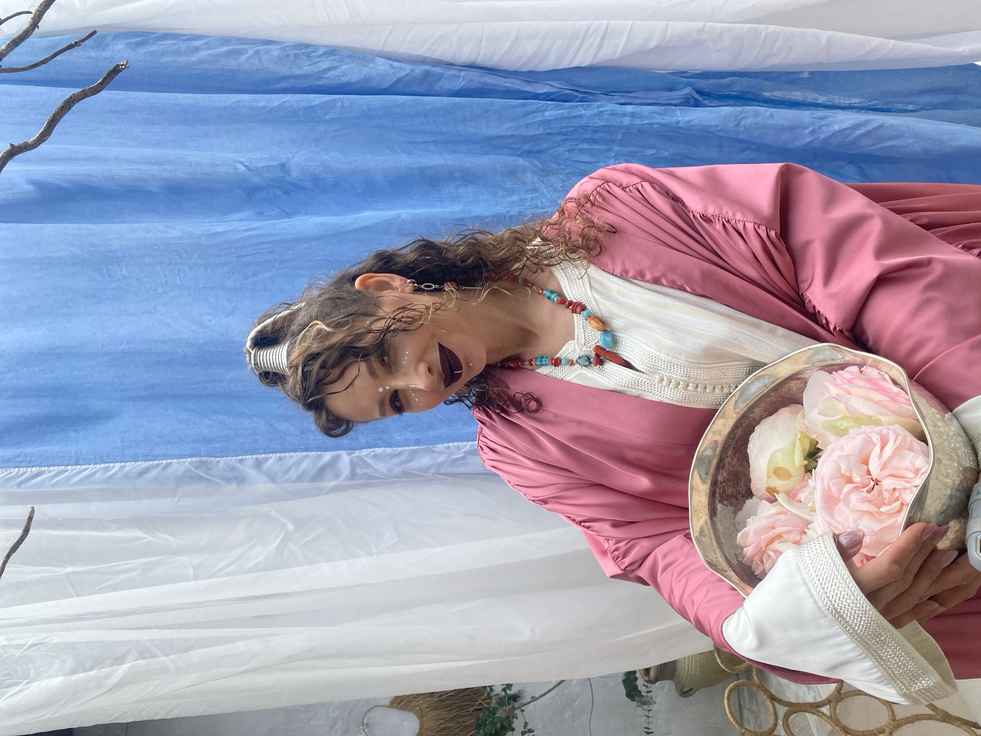 A woman with curly brown hair and elaborate makeup, including white dots on her face and dark lipstick, is holding a silver bowl filled with light pink roses. She is wearing a pink robe over a white garment with intricate detailing, and a colorful beaded necklace. She is positioned in front of a backdrop composed of white and blue draped fabric, with a bare tree branch extending from the top left corner. To the left of the woman, a potted plant and a woven object are visible. The scene appears to be an outdoor setting, possibly a garden or patio, and is located in the city of Sochi, Russia.