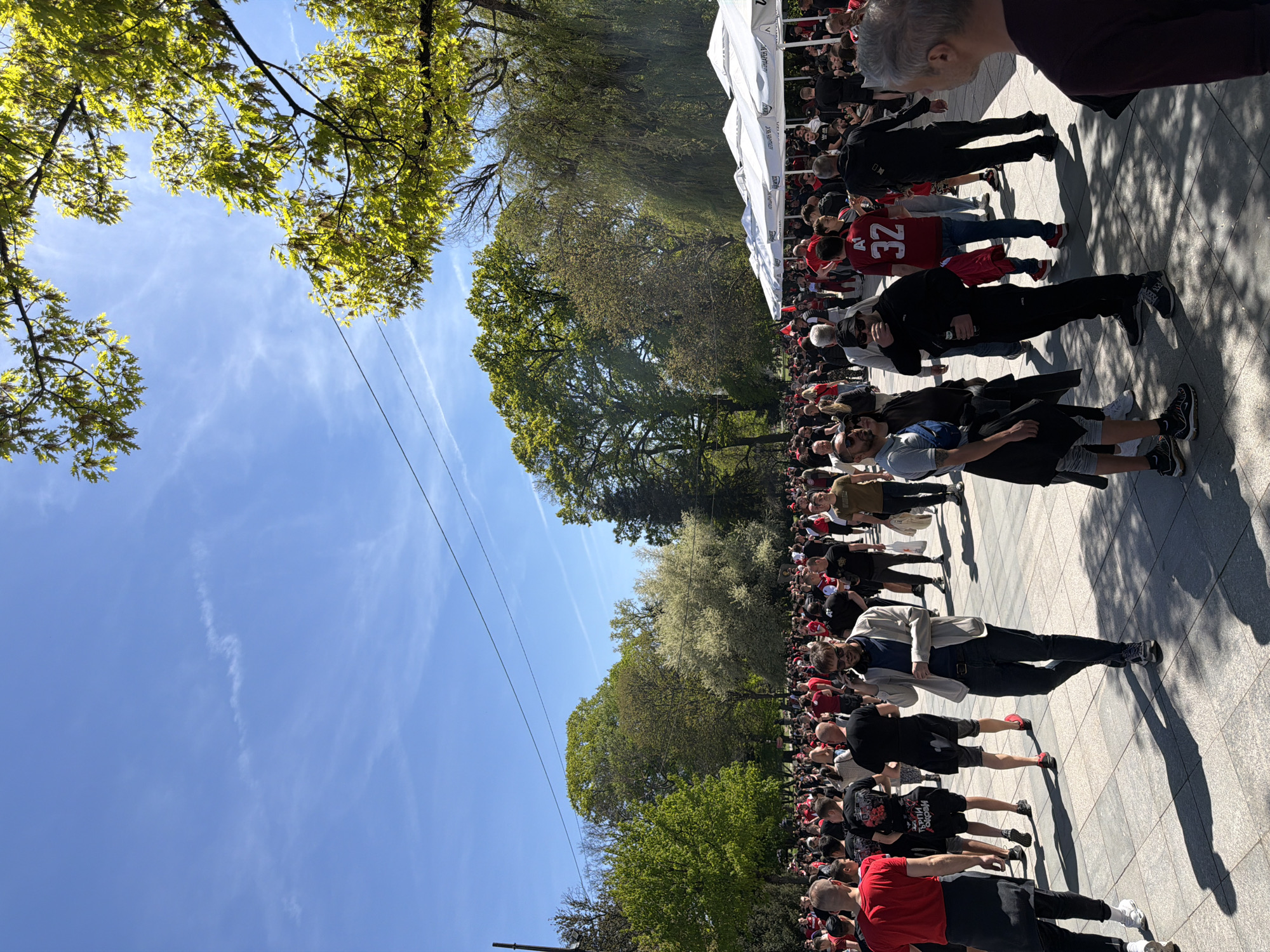 The image shows a large crowd of people, seemingly a gathering or parade, in Sofia, Bulgaria. Many attendees are wearing red and black clothing. Some are wearing jerseys with numbers such as "32" visible. The crowd is situated on a paved area, likely a square or wide sidewalk.
To the left of the crowd, trees are visible, suggesting a park or green space adjacent to the paved area. Overhead, utility wires are present, crossing the sky. Tents or canopies are erected at the edge of the crowd.
The sky is clear and blue, indicating a sunny day. There is a general sense of activity and social interaction within the crowd. The overall impression is that of an organized event or gathering.