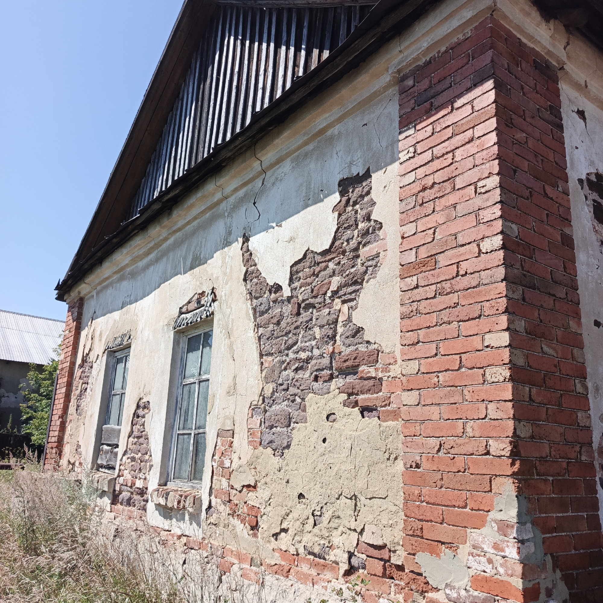 The image shows a dilapidated brick building with crumbling plaster, suggesting it is abandoned or in severe disrepair. The building has a steeply pitched roof covered with dark, corrugated material, possibly metal or slate. The walls are constructed of red brick, much of which is exposed due to the missing plaster. There are two windows visible, with simple frames and glass panes, appearing old and possibly broken. Weeds and tall grass grow around the base of the building. The sky is clear and blue. The context provided is that this building is located in the city of Tugustomir, Russia. The scene depicts an outdoor environment on a sunny day. There are no people or animals visible in the image.