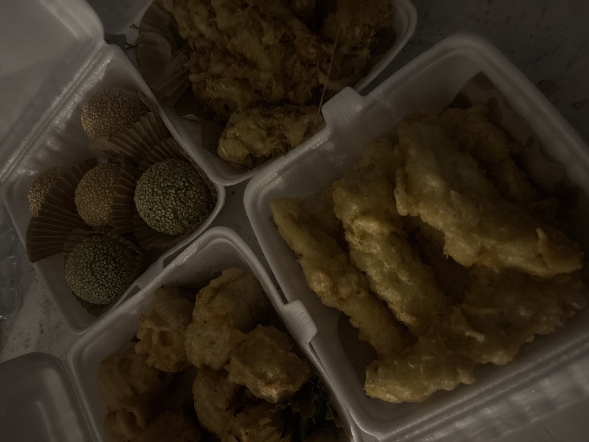 The image captures an overhead view of multiple white foam take-out containers filled with various fried snacks, presented under dim, warm lighting. The overall scene suggests a casual meal or snack delivery, possibly at night or in a poorly lit indoor setting.
In the upper left container, there are several round, spherical pastries coated in sesame seeds, possibly Onde-onde or similar. Some appear golden-brown, covered in white sesame seeds, while others have a green hue, also covered in white sesame seeds. Each pastry is nestled in a fluted paper cup.
Adjacent to this, in the upper right container, is a collection of irregularly shaped, golden-brown fried items, possibly fried tempeh, tofu, or other fritters, appearing somewhat clumpy.
The largest container, occupying the right side of the frame, holds numerous elongated, golden-brown fried items. These strongly resemble fried banana fritters (Pisang Goreng) due to their shape and characteristic fried batter coating.
A fourth container, visible in the lower left, also contains various irregularly shaped fried pieces, similar to those in the upper right container, suggesting another type of fritter.
The food items generally exhibit a golden-brown color, indicative of deep frying. No people are visible, nor is any specific activity taking place other than the presentation of the prepared food. There is no visible text on any of the containers or food items. The low light conditions contribute to a somewhat muted and warm color palette.