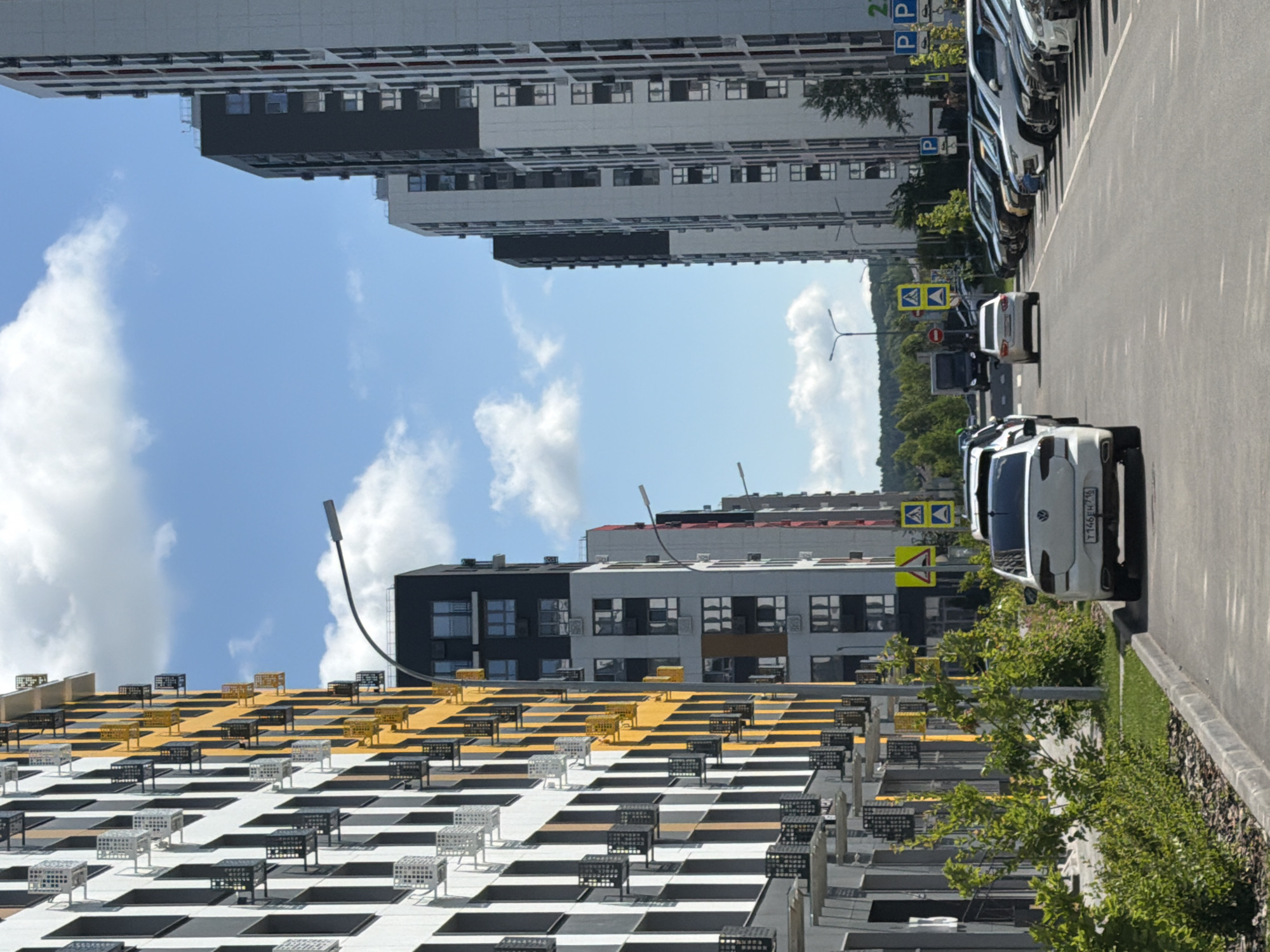 A street scene is depicted with modern residential buildings lining both sides. On the left, a tall building features a black and white facade with yellow accents and numerous balconies, each fitted with air conditioning units. On the right, another modern building displays a predominantly white exterior with black and red details. The street is paved and a line of parked cars is visible on the right side, with one white car in the foreground. Road signs, including pedestrian crossing and parking symbols, are present. Green trees and bushes line the sidewalk on the left. The sky is blue with scattered white clouds. The overall impression is of a new urban development.