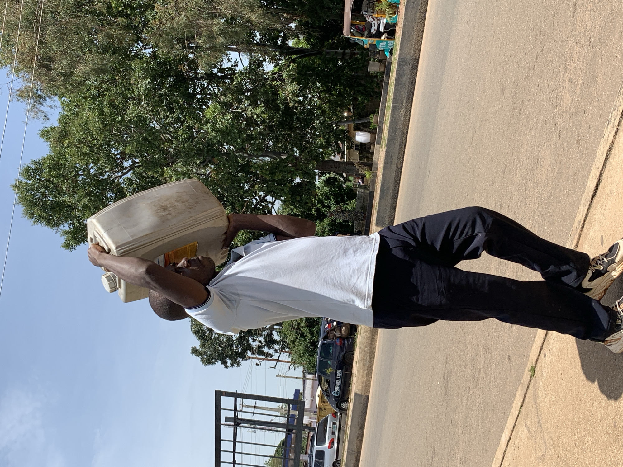 A man walks on a paved sidewalk next to a road, carrying a large, beige plastic jerrycan filled with liquid on his shoulders. He is wearing a light blue short-sleeved polo shirt and dark blue trousers. The jerrycan has a white cap and some yellow labeling visible. In the background, there is a road with a few vehicles, including a dark minivan with "SIFAWA LINE" written on it. Trees line the side of the road, and power lines are visible overhead. The scene appears to be in a city, with buildings and infrastructure visible in the distance. The man is walking towards the right side of the frame, and the sun is casting a shadow from him onto the sidewalk. The overall impression is one of a person transporting a load through an urban environment on a sunny day. The location is identified as the city of Jos, Nigeria.