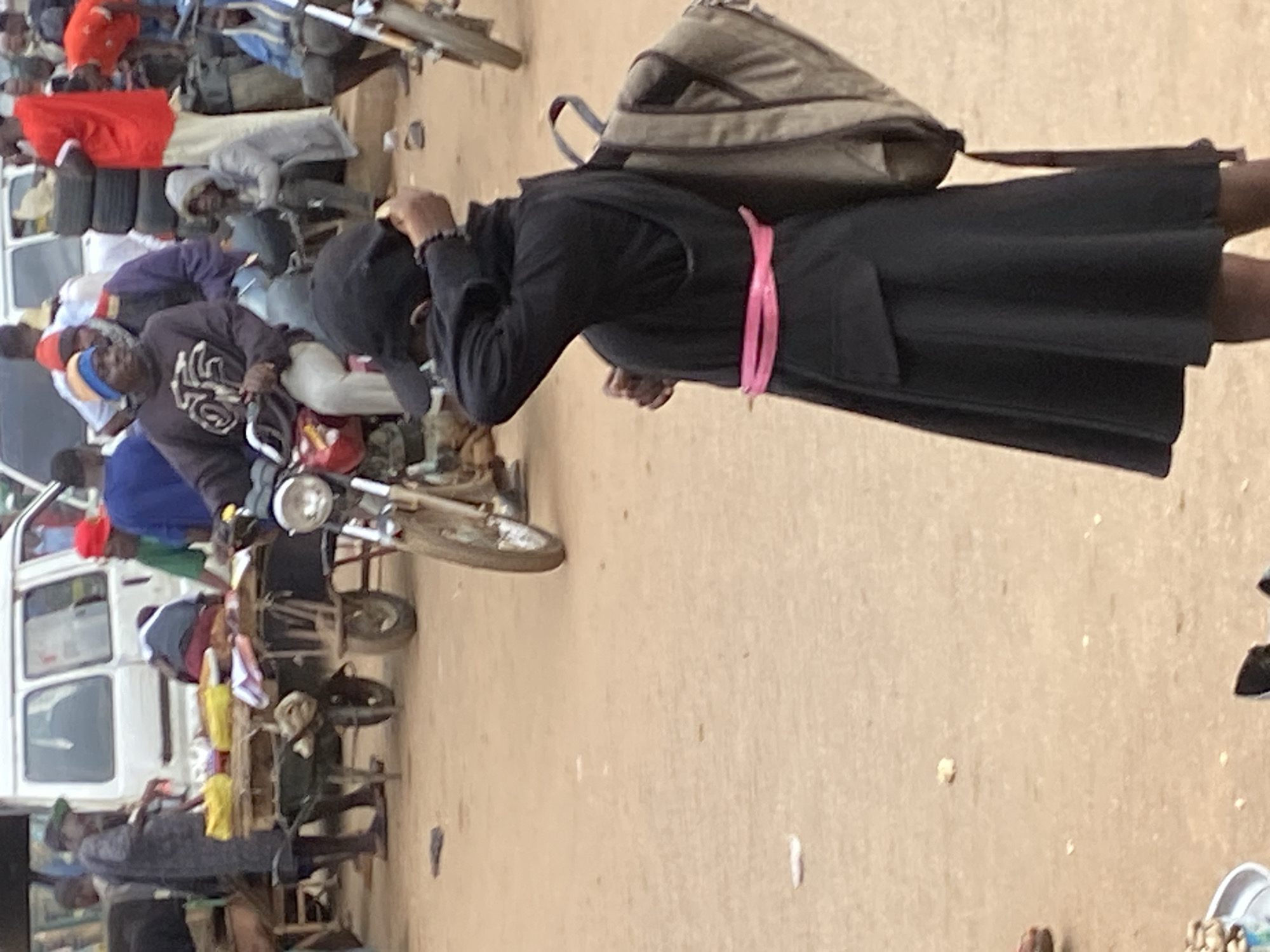 In Bukuru, Nigeria, a busy street scene unfolds. A young woman, clad in a black abaya with a pink sash and a black hijab, stands with her back to the camera, her hand reaching up to adjust her head covering. She carries a beige backpack. In the background, a man in a blue sweater and jeans rides a motorcycle, navigating through the bustling market. Various stalls are laden with goods, and people mill about, engaged in commerce and daily activities. The environment is sandy and dusty, typical of a Nigerian street market.