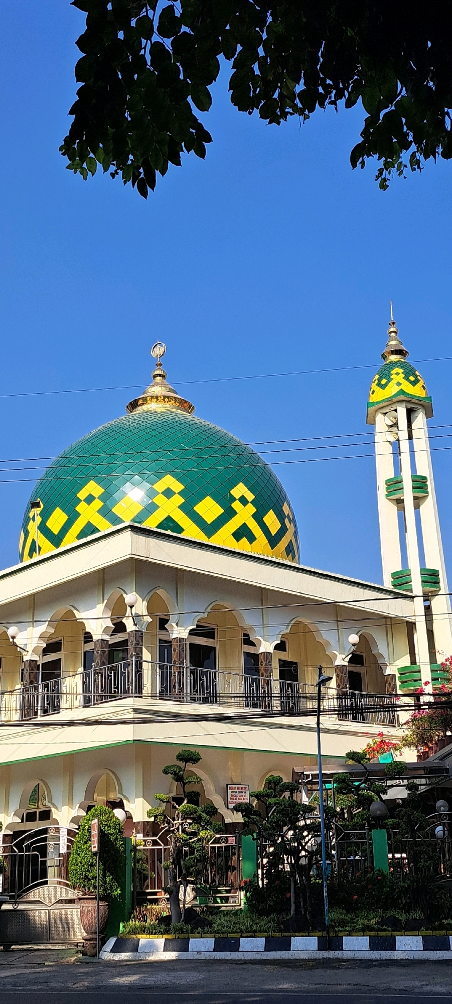 A daytime, eye-level, vertical, full shot captures the exterior of a mosque in Karang Jati, Indonesia. The mosque features a large, green and yellow patterned dome topped with a crescent moon finial, and a tall, slender minaret with a similar patterned top. The building is painted in a light yellow color and has arched windows and balconies with railings. Several power lines crisscross the sky above the structure. In the foreground, there is a neatly landscaped area with manicured trees and bushes, bordered by a decorative black and white striped curb and a road. The sky is a clear, bright blue. There are no people visible in the image.