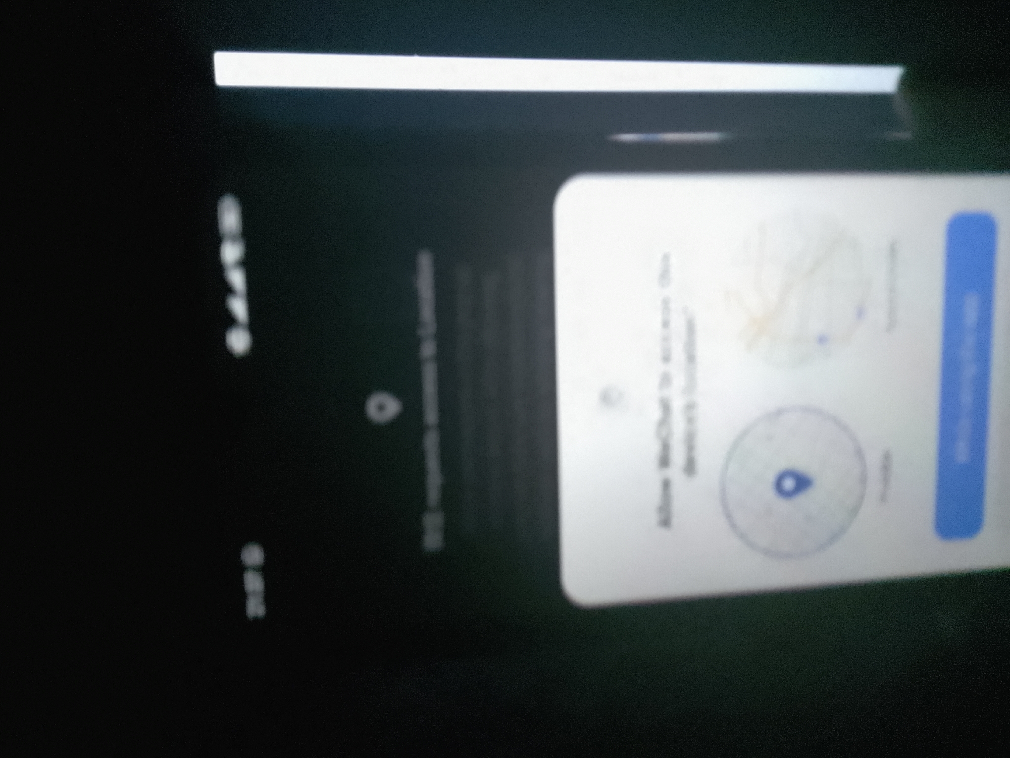 The image shows a smartphone screen displaying a WeChat location permission request. The request asks to "Allow WeChat to access this device's location." There are two circular icons, one with a map-like graphic and another with a blue map marker. A blue button with the text "Allow" is visible on the right side of the screen. The phone is held at an angle, and the background is dark. The overall scene is consistent with a user interacting with a mobile application. There is no identifiable information or context related to the city of Azume, Nigeria.