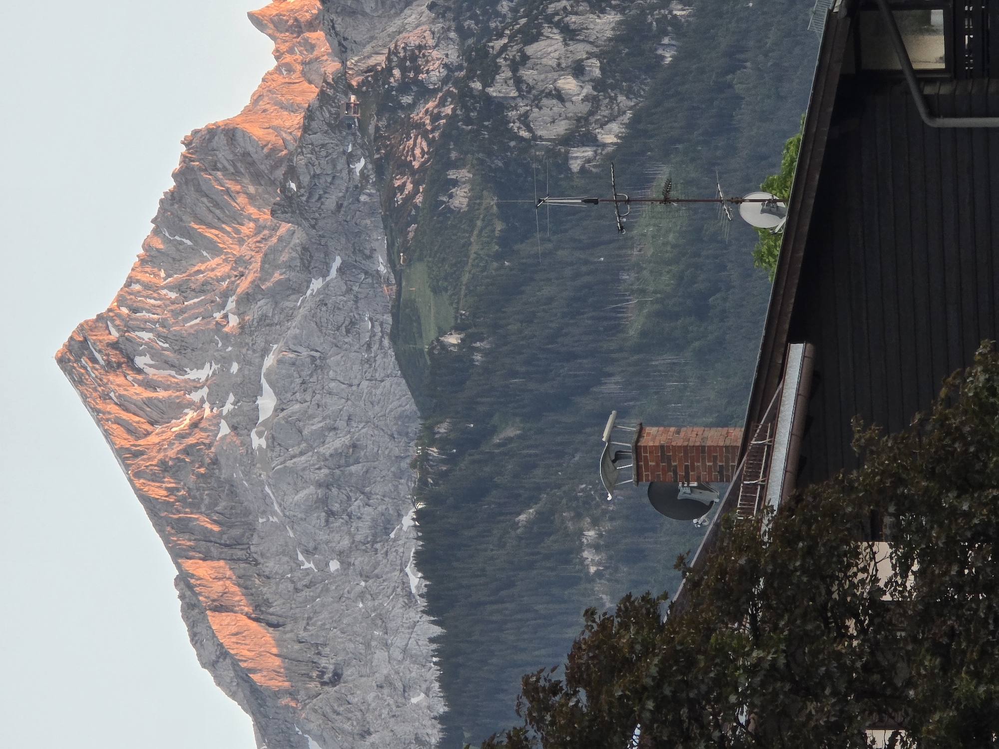 This is a photograph of the Zugspitze mountain in Garmisch-Partenkirchen, Germany, taken at sunset. The foreground shows the roof of a dark-colored wooden house with a brick chimney, a satellite dish, and an antenna. A dark tree with large leaves is partially visible on the left side of the frame. The middle ground features a dense, dark green forest. The background is dominated by the rugged, rocky slopes of the Zugspitze, illuminated by the warm, orange light of the setting sun, with patches of snow visible on its peaks. The sky is a clear, pale blue.
