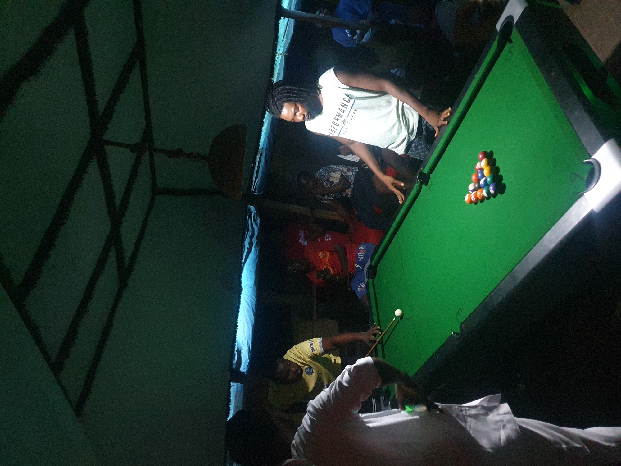 This indoor scene depicts several individuals engaged in a game of pool, likely in an establishment in Lokoja, Nigeria. The image is oriented at an angle, giving a tilted perspective.
The primary subjects are a man with dreadlocks and a beard, wearing a light-colored t-shirt with the word "PERFORMANCE" prominently displayed, standing at one end of a green pool table. He is looking down at the table, seemingly observing the game or preparing for a shot. At the opposite end of the table, another man in a yellow t-shirt with a blue collar is actively playing, leaning over the table with a pool cue, poised to strike the cue ball. Other individuals are partially visible in the background, suggesting a small gathering. One person in a white shirt is in the foreground, partly obscured, while others in red and blue shirts are seen further back.
The main object is a standard green-felt pool table. At one end, a full rack of billiard balls, including solid and striped balls, the black 8-ball, and a variety of colors like red, yellow, and blue, is neatly arranged. A white cue ball is positioned at the other end of the table. Pool cues are also visible. The setting appears to be dimly lit, likely evening or night, with focused artificial lighting illuminating the pool table. Overhead, a structure of dark, possibly wooden beams or supports, is visible against a dark green ceiling, contributing to a rustic or informal atmosphere. No specific weather conditions are discernible as it's an indoor shot. The emotions of the participants appear to be focused on the game.
