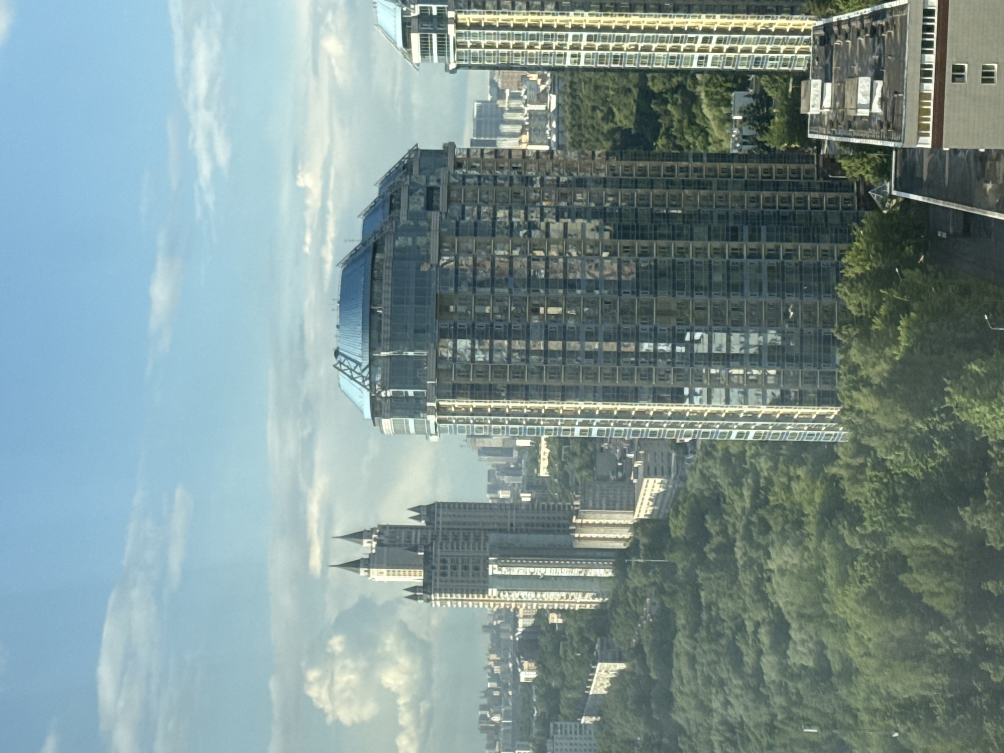 The image captures a high-angle view of a cityscape, with a large, modern glass-clad building dominating the frame. Other buildings, including one with turret-like structures, can be seen nestled amongst lush green trees and cityscape. The sky is partially cloudy, and the general environment appears to be urban with a significant presence of greenery. It is difficult to determine the location specifically as the city of Khimki, Russia.