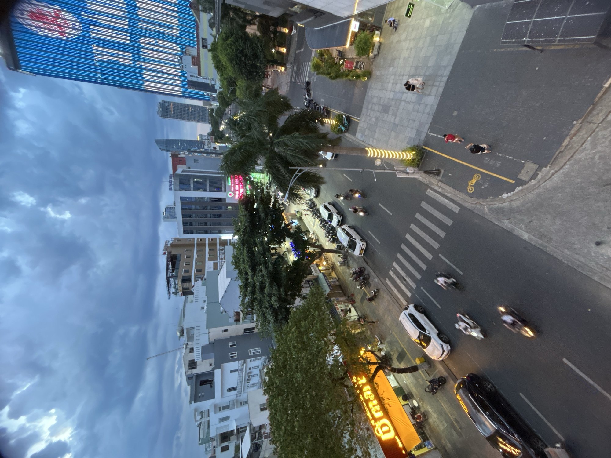 This is an aerial, slightly fisheye lens perspective shot looking down onto a street in Đà Nẵng, Vietnam, at dusk. The sky is filled with dramatic, dark clouds.
On the right side of the image is a tall, modern building with a facade covered in bright blue and white lights that form a pattern and what appears to be a red logo. In the foreground, on the right, is a paved walkway with a few people sitting and standing, and a dark-colored structure that could be part of a building or a large piece of equipment.
The street itself is busy with traffic. There are several cars and many motorcycles. On the left side of the street, there is a building with a bright orange illuminated sign that says "D'mart" in yellow lettering. There are trees lining the street, some with decorative yellow lights wrapped around their trunks. A palm tree stands prominently in the middle of the scene. Pedestrians are visible on the sidewalks and crossing the street at a crosswalk. A yellow motorcycle symbol is painted on the pavement near the crosswalk.