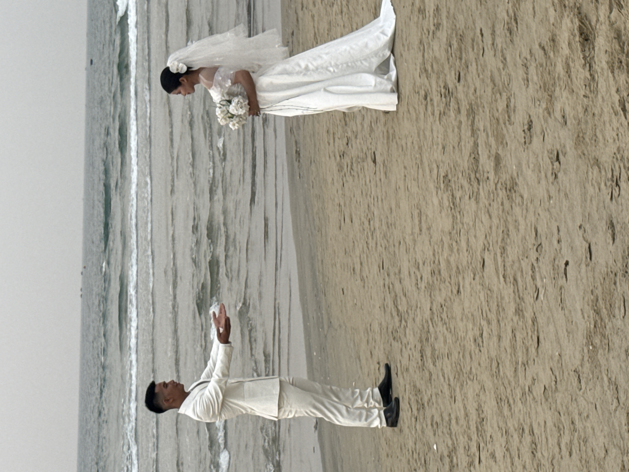 The image shows a bride and groom on a beach. The bride is wearing a white wedding dress and veil and holding a bouquet of white flowers. The groom is wearing a white suit with black shoes and has his hands raised. They are standing on a sandy beach with ocean waves visible in the background. The location is likely My Khe Beach in Đà Nẵng, Vietnam, due to the appearance of the sand and the ocean.