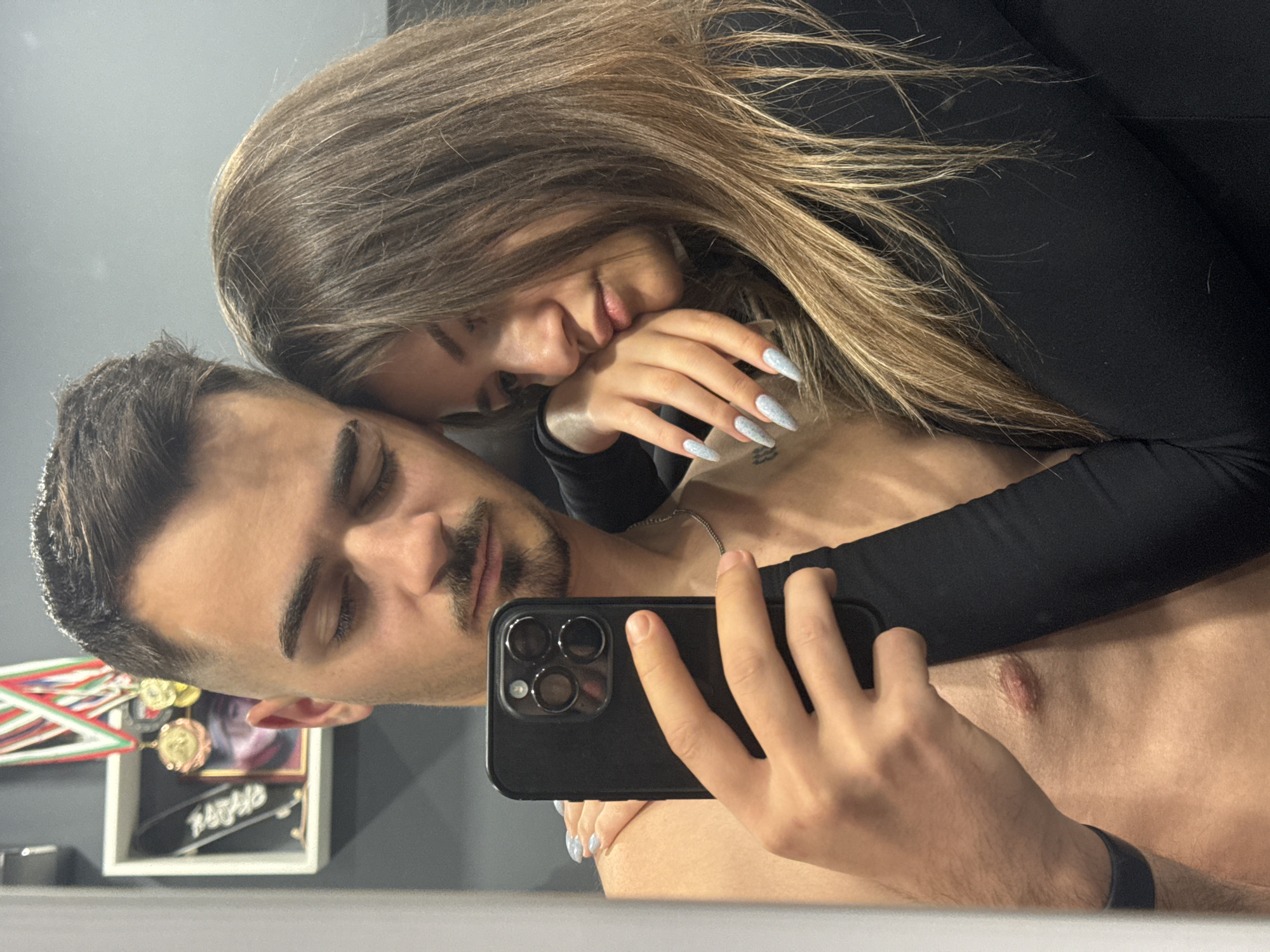 A selfie shows a shirtless man with short, dark hair and a mustache holding a black smartphone. A woman with long, light brown hair and long, grey nails is leaning on the man's shoulder. The woman is wearing a long-sleeved black shirt. A framed photograph, medals, and a skateboard are visible on a shelf in the background. The location is potentially Sofia, Bulgaria, due to the presence of medals in the background with the Bulgarian flag colors. The man is possibly taking the selfie.