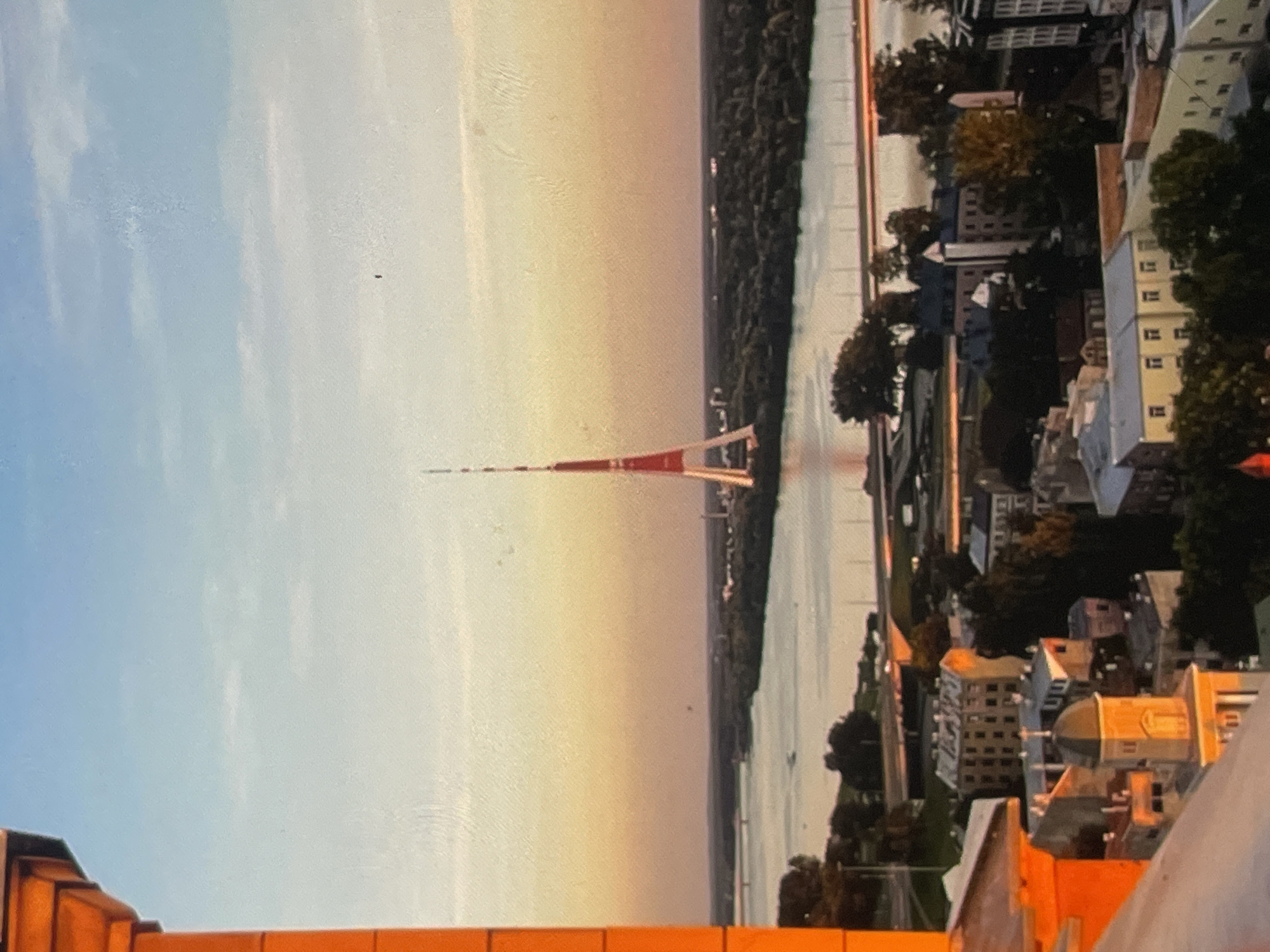 The image captures a panoramic view of a city at sunset. Dominating the skyline is a tall, slender tower with a red and white lattice structure, resembling a television or radio mast. A wide river flows through the city, with bridges spanning across it. The foreground shows a cluster of buildings with various architectural styles, including a prominent building with a dome topped by a cross, suggesting a religious structure. The sky is a gradient of warm colors, transitioning from orange and yellow near the horizon to blue overhead, with a few wispy clouds. Trees and greenery are interspersed throughout the urban landscape, particularly along the riverbanks and surrounding the buildings. The overall impression is one of a serene, urban setting during the golden hour.
The requested location in the city of Harmanli, Bulgaria, cannot be confirmed from the visual information present in the image. There are no identifiable landmarks or textual cues within the image that specifically link it to Harmanli, Bulgaria.