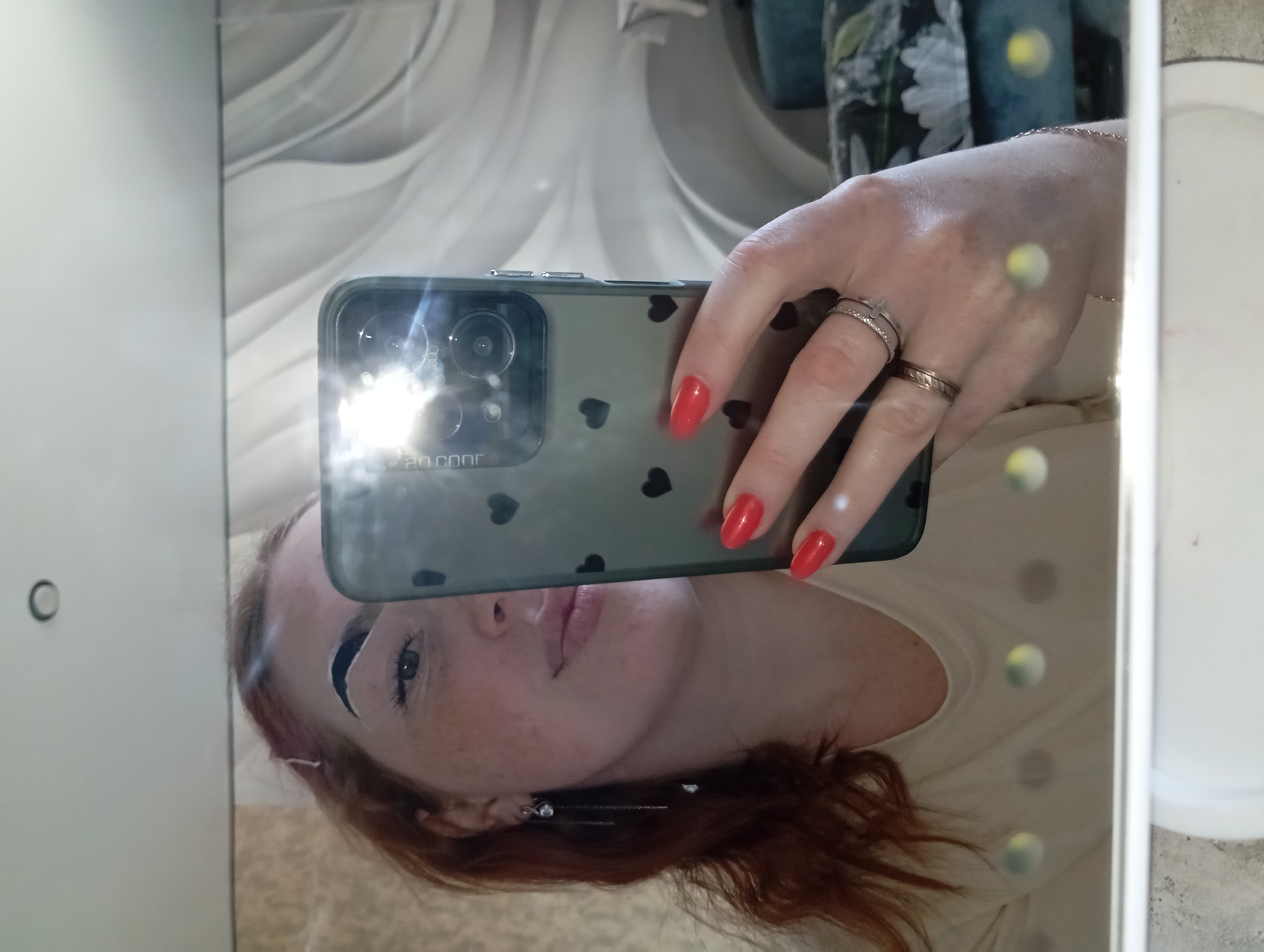A red-haired woman is taking a selfie in a mirror. The phone she is using is a dark color with small black hearts on the back, and has 3 camera lenses, two rings are seen on her hand with red painted fingernails. The background includes a marble-patterned wall and a floral design. The location is unknown and cannot be determined from the image.