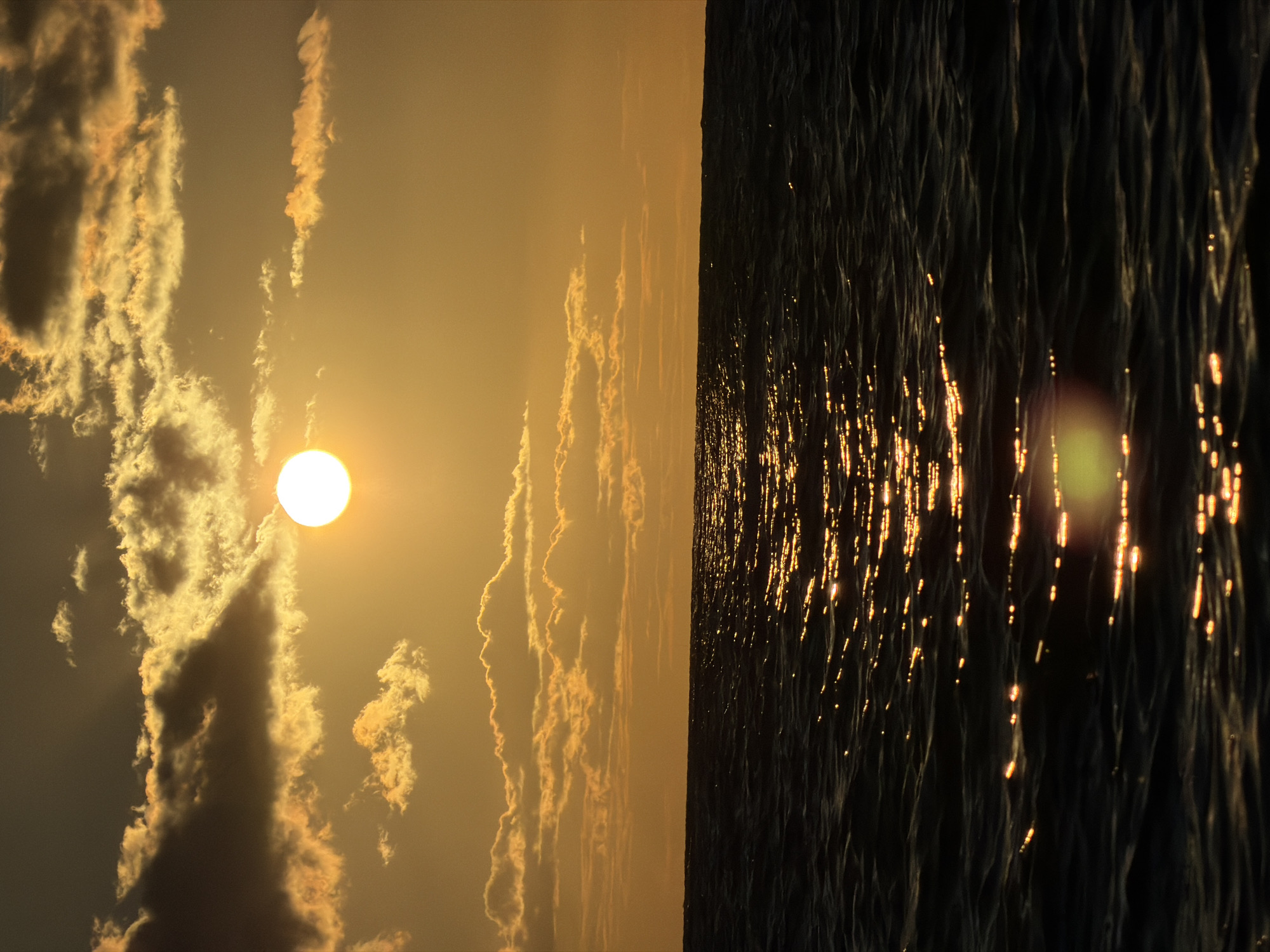 The image depicts a sunset over the ocean. The sun, a bright orb, is positioned in the upper center of the frame, partially obscured by golden clouds. Rays of light emanate from the sun, illuminating the sky with warm orange and yellow hues. The ocean below is dark and rippling, with the sun's reflection creating streaks of light across its surface. The horizon line is sharp, separating the sky from the sea. There are no people or man-made objects visible in the image, suggesting a natural, serene environment. The location is identified as Baan Bo Phut, Thailand, a coastal area known for its scenic sunsets.