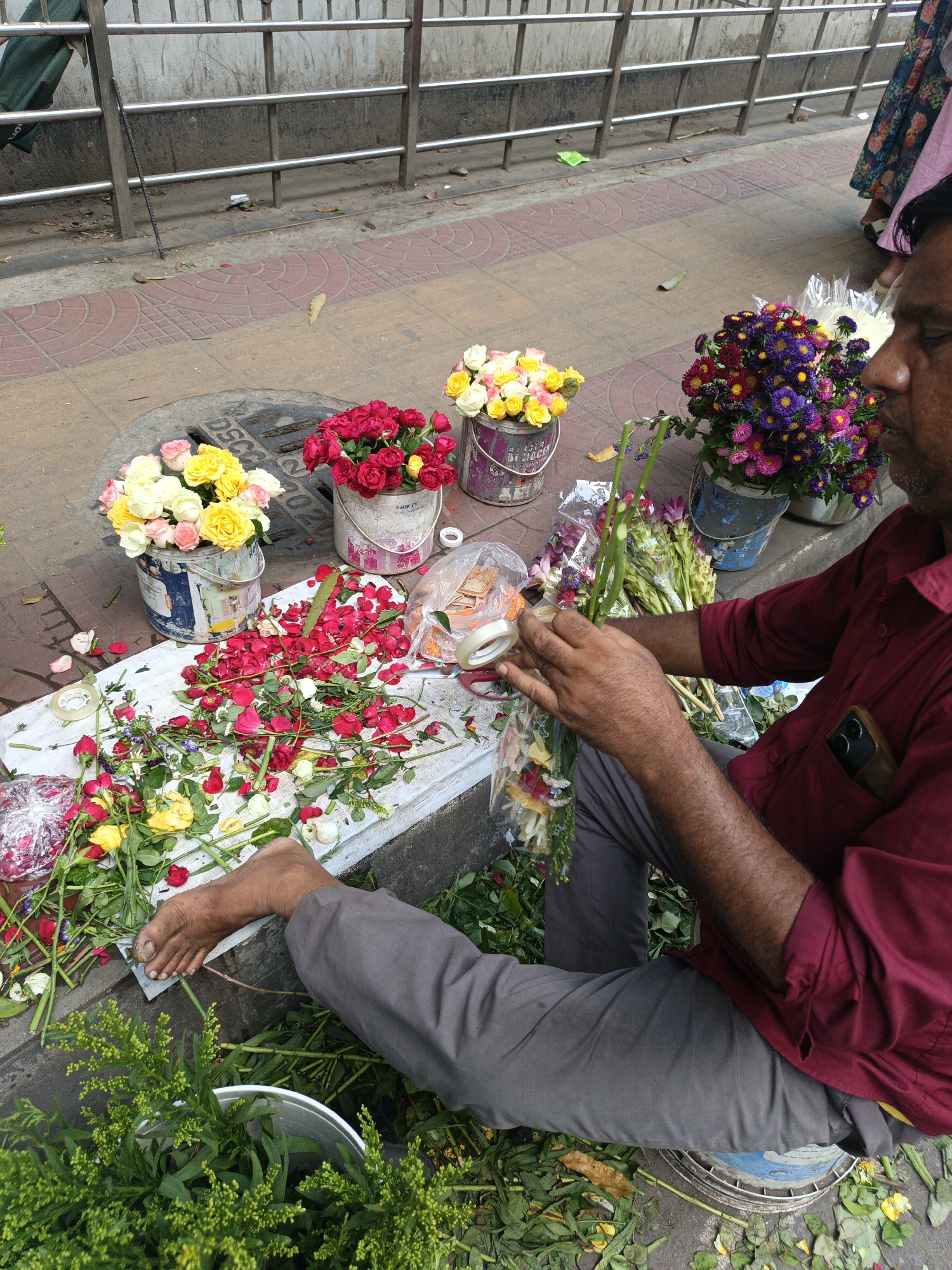 An Indian flower vendor sits cross-legged on the pavement on a city street in Dhaka, Bangladesh. The man is light-skinned with dark, thinning hair and a close-cropped beard. He is wearing a maroon long-sleeved shirt and gray pants, and he has a cell phone in his shirt pocket. The vendor is preparing flower bouquets, using clear tape. Buckets of roses and other flowers are arranged around him on the pavement. One bucket contains yellow and white roses, another contains red roses, and a third bucket contains a mix of colorful flowers. Scattered on a white surface next to the buckets are loose flower petals, stems, and leaves. The flowers are mainly red and white roses and purple and yellow chrysanthemum-like flowers. The vendor is sitting on a blue-and-white bucket. Metal railings line the back of the pavement, and the pavement itself is made of red and yellow brick.