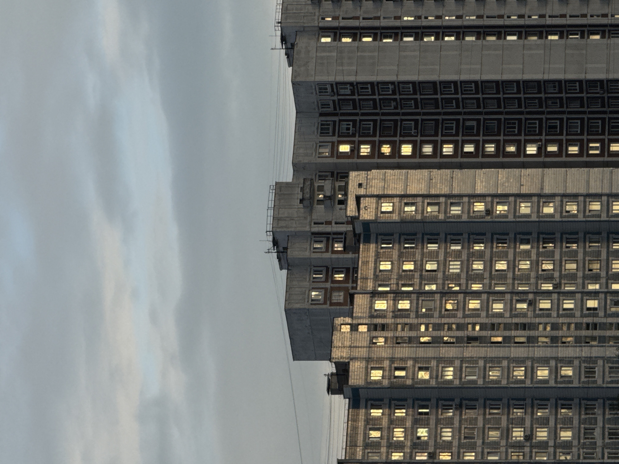 The image shows a close-up view of the upper floors of several tall, grey residential buildings in Moscow, Russia. The buildings are constructed in a modular, prefabricated style, common in Soviet-era urban development. The windows of the buildings are illuminated with a warm, yellow light, suggesting that it is either dusk or dawn, and the lights are on inside the apartments. The sky above is overcast with grey and pale blue clouds. Power lines are visible strung between the buildings in the upper portion of the frame. There are no people or visible activity outside of the illuminated windows. The scene is characterized by the repetitive geometric patterns of the building facades and windows.
