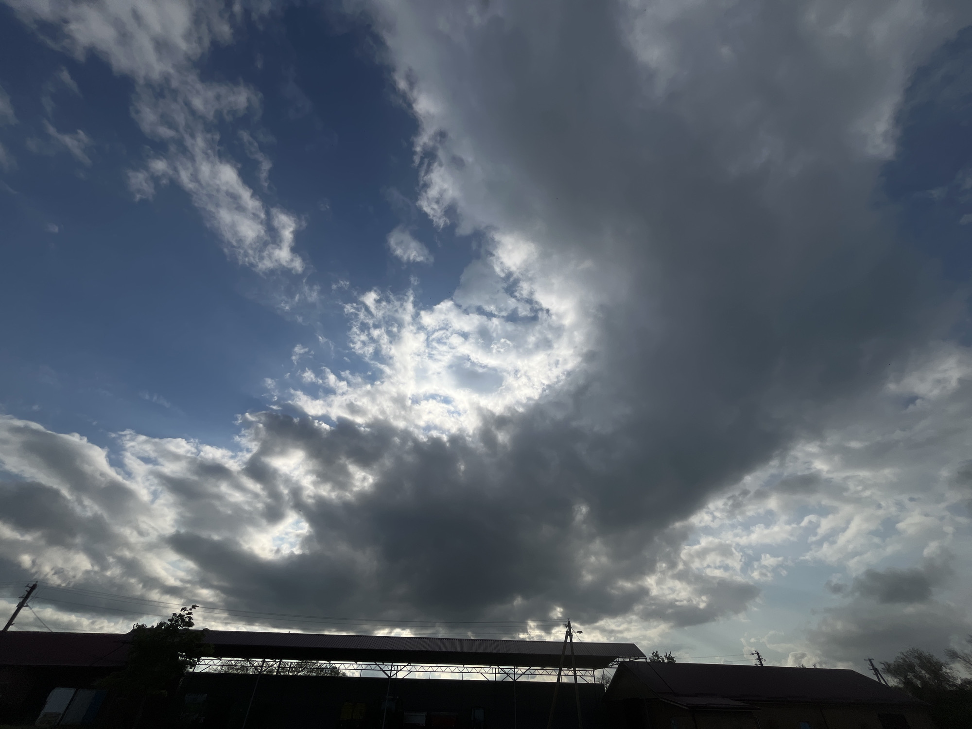 The image shows a view of the sky in Novopokrovskaya, Russia. The sky is partially covered with clouds, with patches of blue sky visible in between. A large, dark cloud formation dominates the right side of the frame. Below the sky, the roofs of several buildings are visible, suggesting a residential or commercial area. A structure with a metal framework is also present.