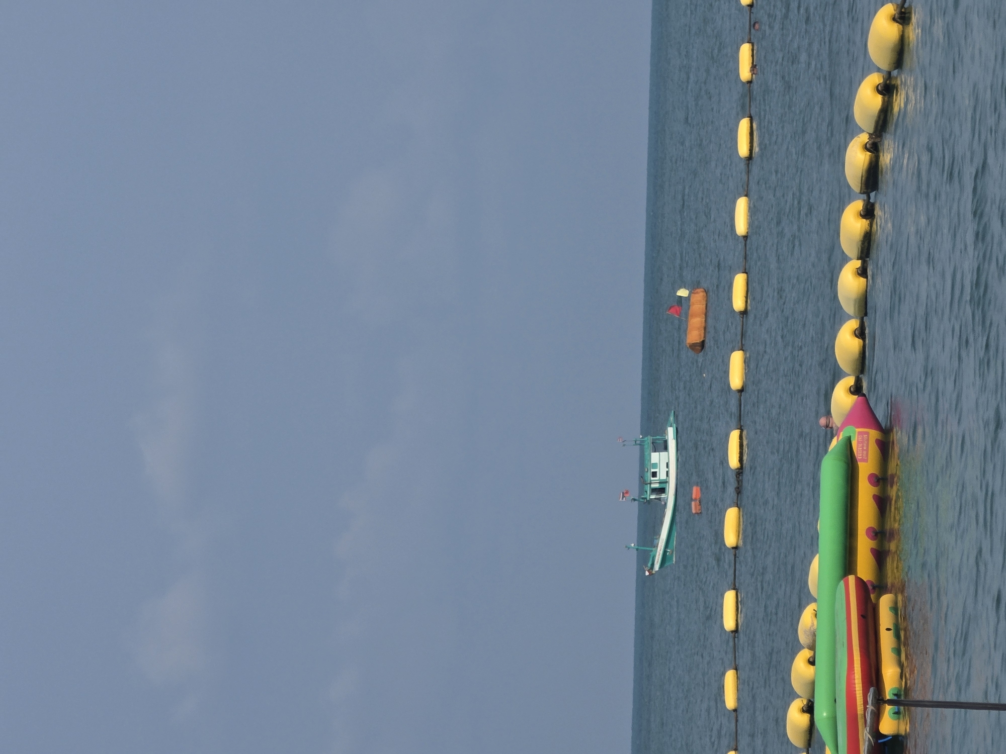 The image depicts a seascape in Pattaya City, Thailand, featuring a body of water under a cloudy sky. Running vertically on the right side of the frame, a chain of yellow buoys marks a zone within the sea. Adjacent to this marked area, inflatable water toys, including a long green tube and a colorful banana boat, float near the buoy line. In the distance, a fishing boat sits on the water. Further away, a smaller, oblong object is visible amidst the sea.