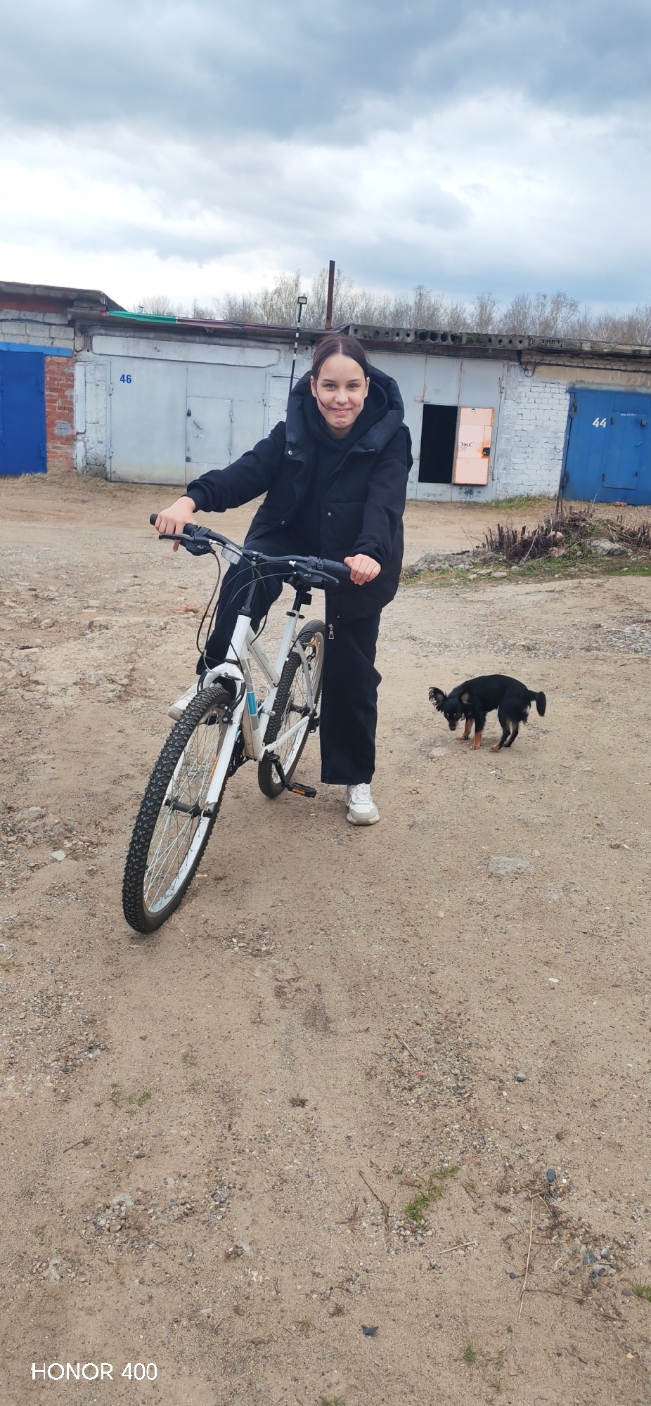 The image depicts a young woman on a bicycle in a location with garage structures, likely in Noviy, Russia. The woman, wearing a black jacket and black pants, is positioned to the left of a white bicycle. A small black and tan dog stands nearby. The garages in the background are numbered 44 and 46, suggesting a structured storage or maintenance area. The ground appears to be dirt or gravel, and the sky is overcast.