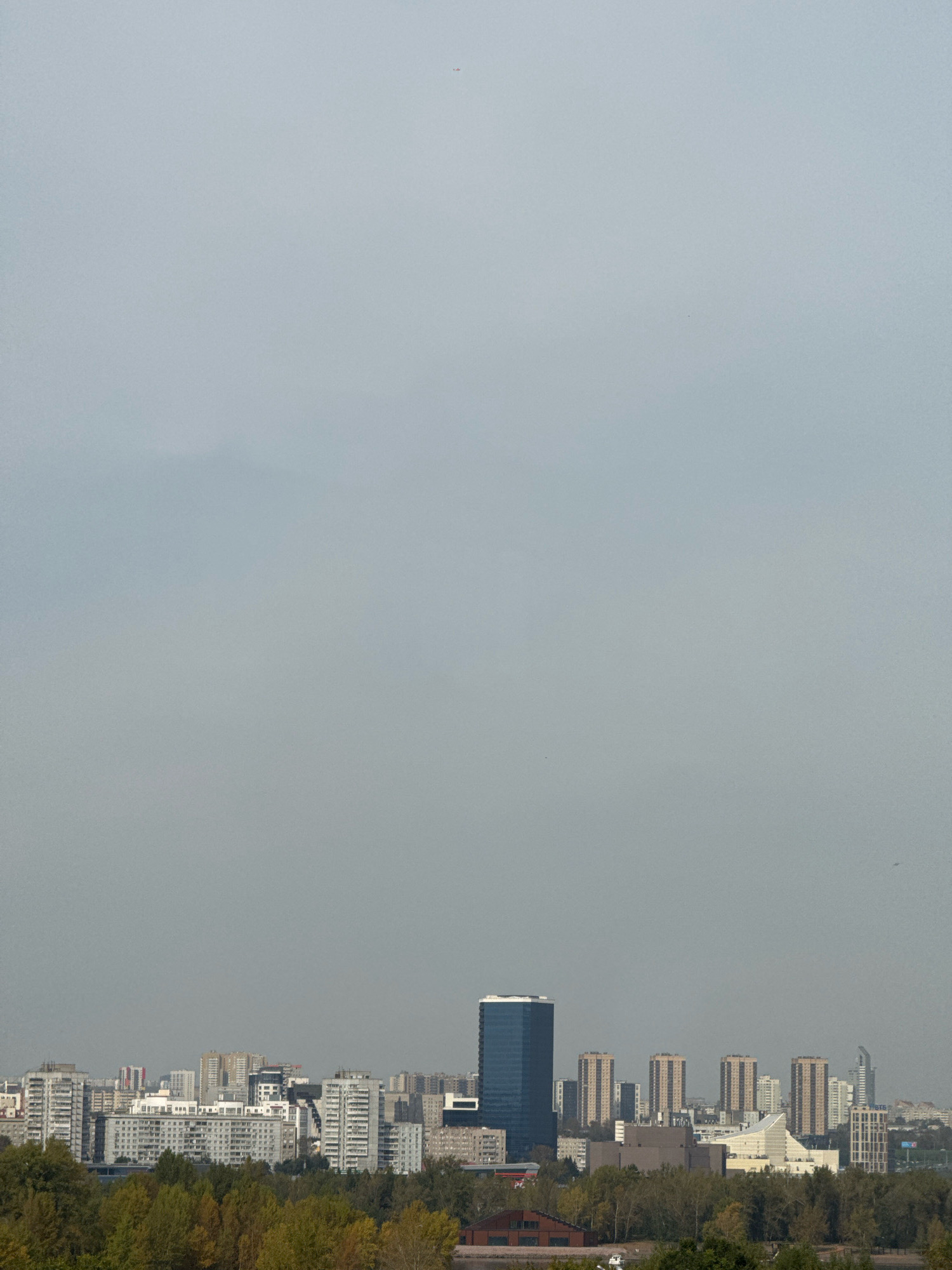 The image depicts a city skyline under a hazy, overcast sky. In the foreground, a dense band of trees with autumnal foliage runs horizontally across the bottom of the frame. Behind the trees, a collection of mid-rise and high-rise buildings forms the urban landscape. A prominent, dark blue skyscraper stands out amongst the other structures. The overall impression is that of a residential and commercial area within a city. Based on the distinctive architecture, specifically the blue skyscraper and the cluster of beige high-rises, this scene is identifiable as the Pokrovsky district in Krasnoyarsk, Russia, overlooking the Yenisei River.