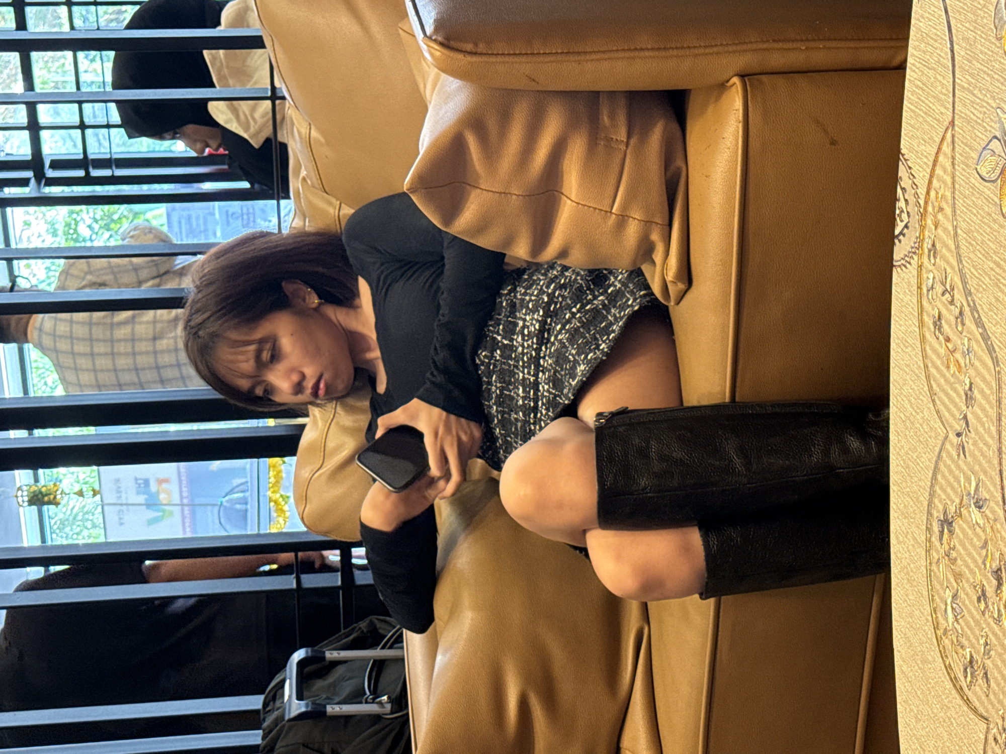 A woman with short brown hair is sitting on a brown leather couch, wearing black knee-high boots, a black long-sleeved shirt, and a grey tweed skirt. She is holding a black smartphone in her left hand. A second woman in a black hijab is visible in the background through a window. Another person is wearing a plaid shirt in the same background. There is a black suitcase with handles and wheels on the floor next to the couch. The location is Kuala Lumpur, Malaysia, and a building with a sign is visible in the background.